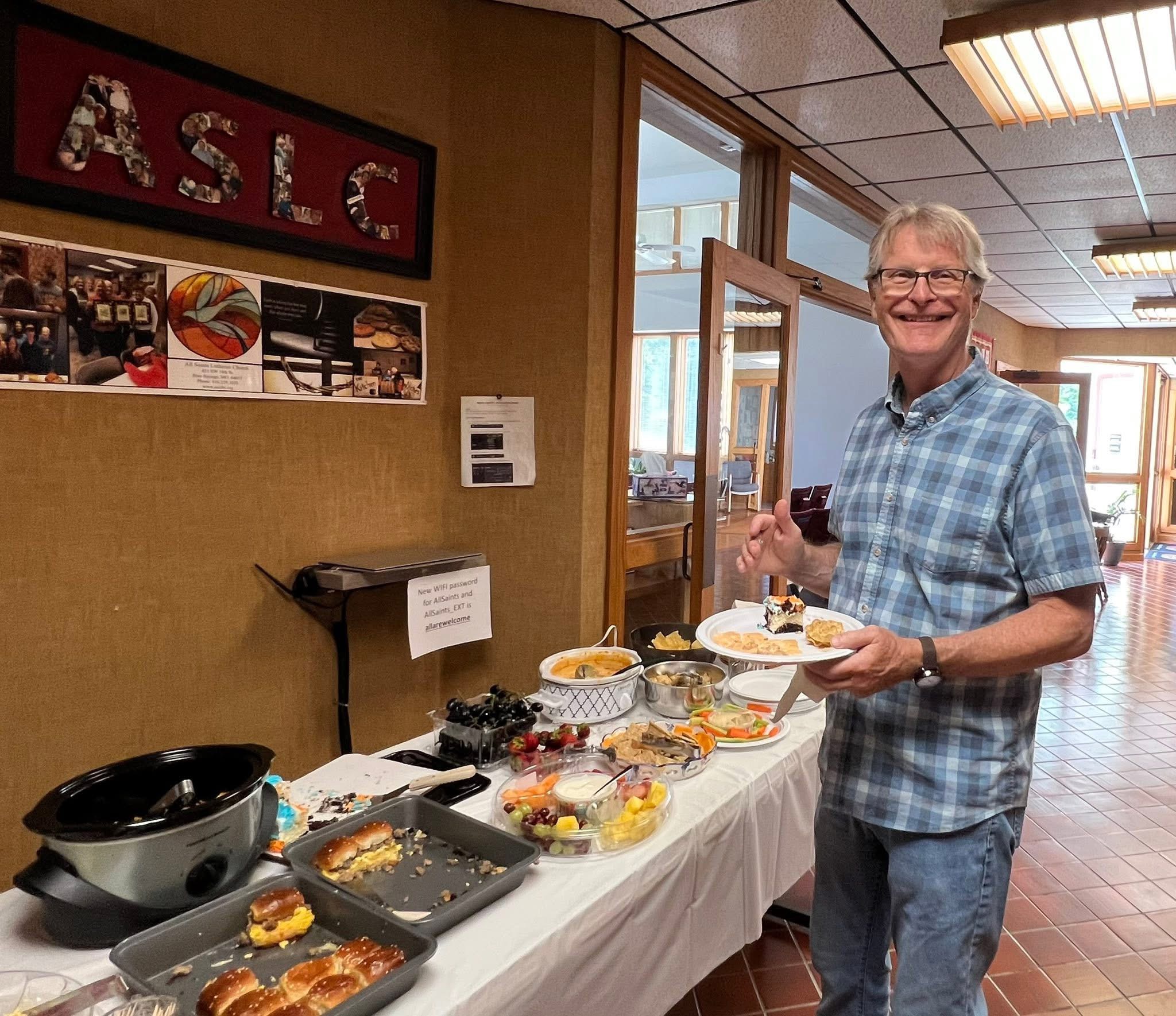 Smiling man in plaid shirt holding plates at buffet table with food, in room with windows and ASLC sign.