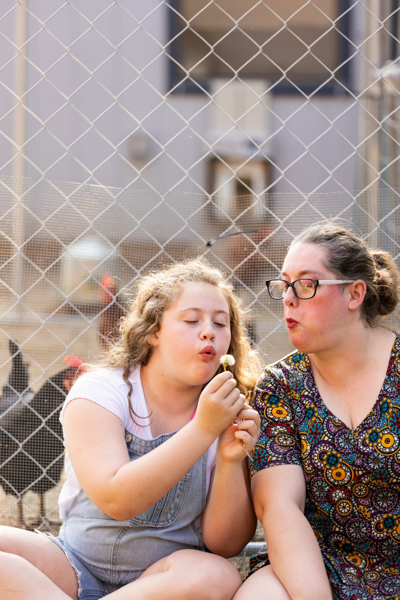 Two girls sit behind a chain-link fence, one holding a dandelion, with a blurred background of buildings.