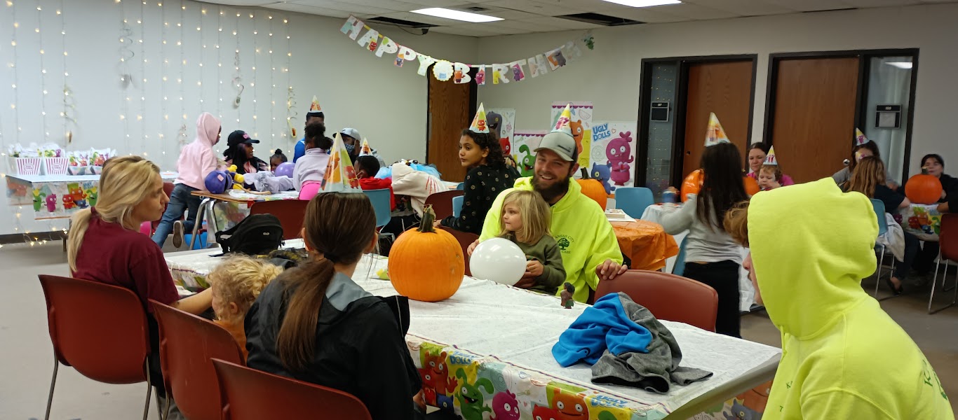 People gathered around tables decorated for a celebration, with balloons and pumpkins, in a decorated room.