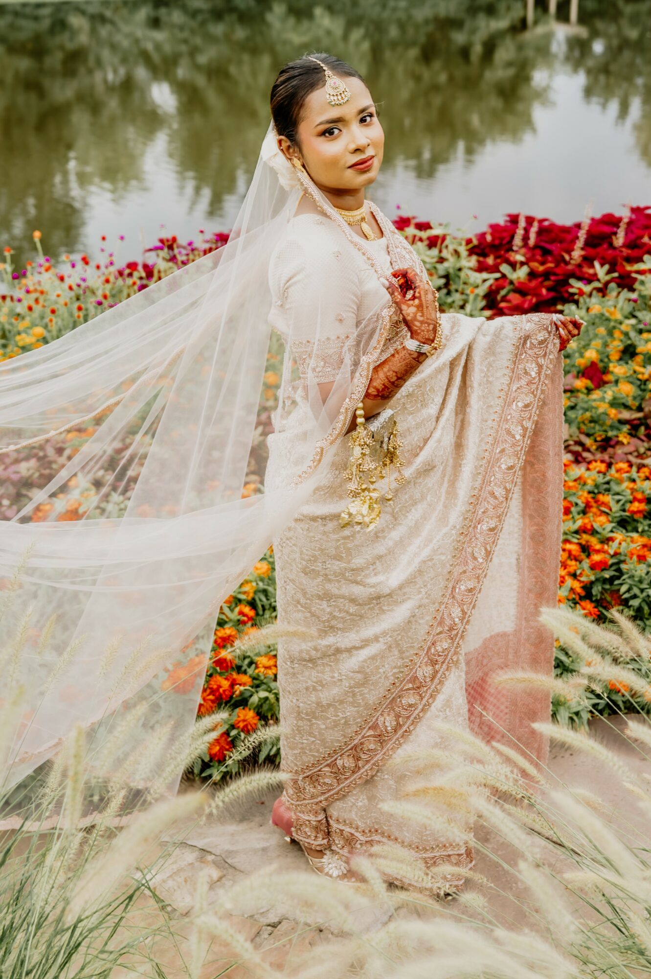 Woman in traditional attire standing in a flower garden near water, holding her dress, with a veil flowing behind her.