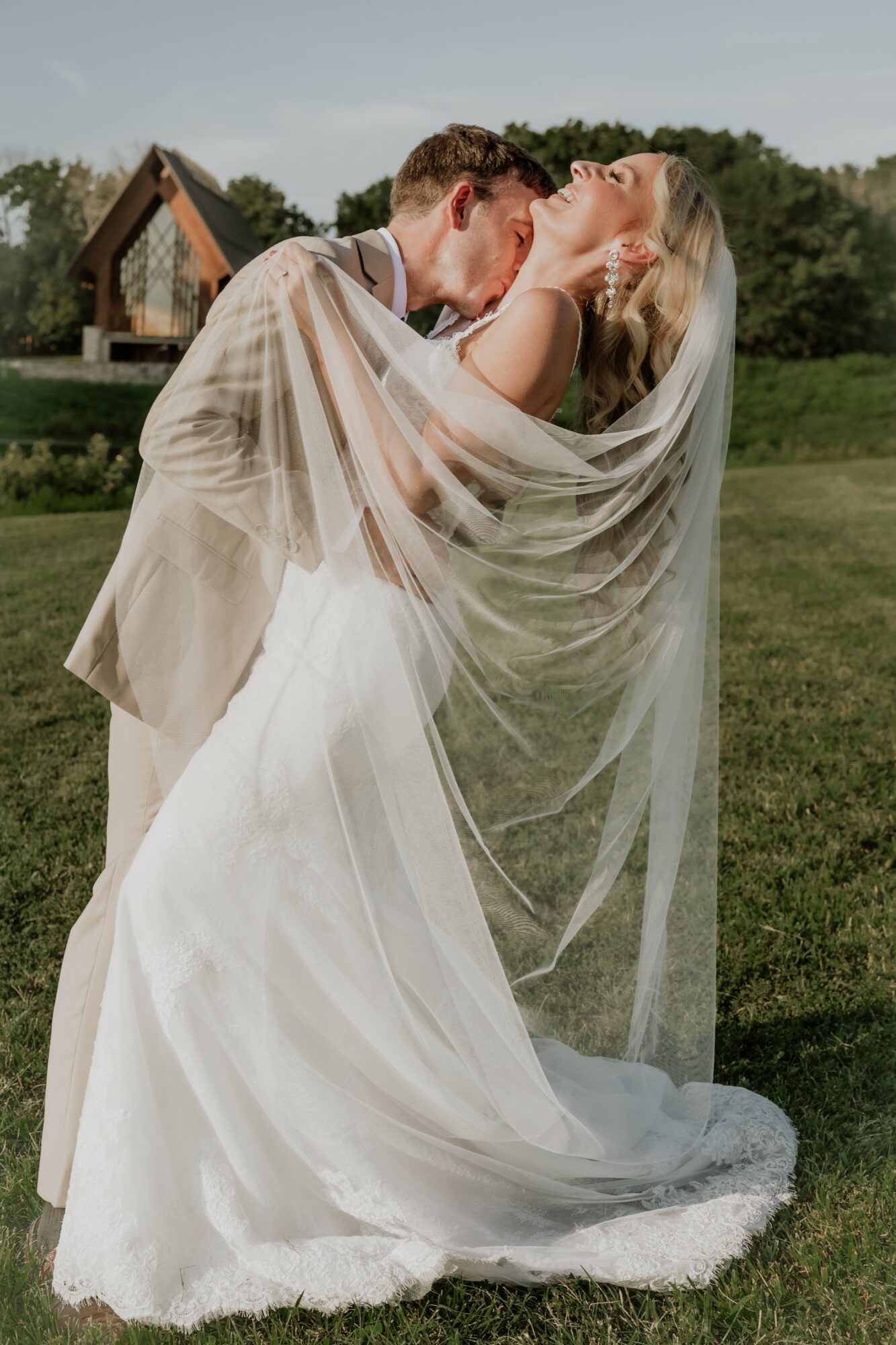A couple in wedding attire outdoors, with the man lifting the woman, who is wearing a veil, in a grassy area.