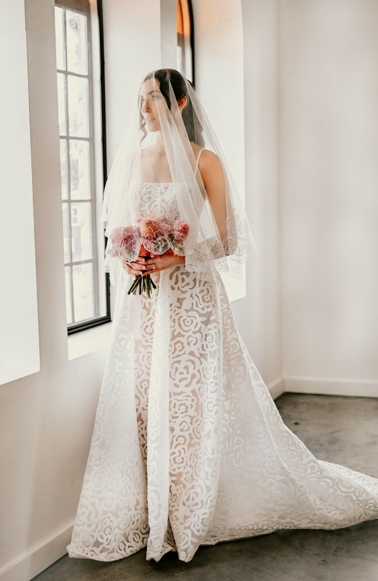 Bride in a lace wedding dress holding a bouquet, standing by a window indoors.