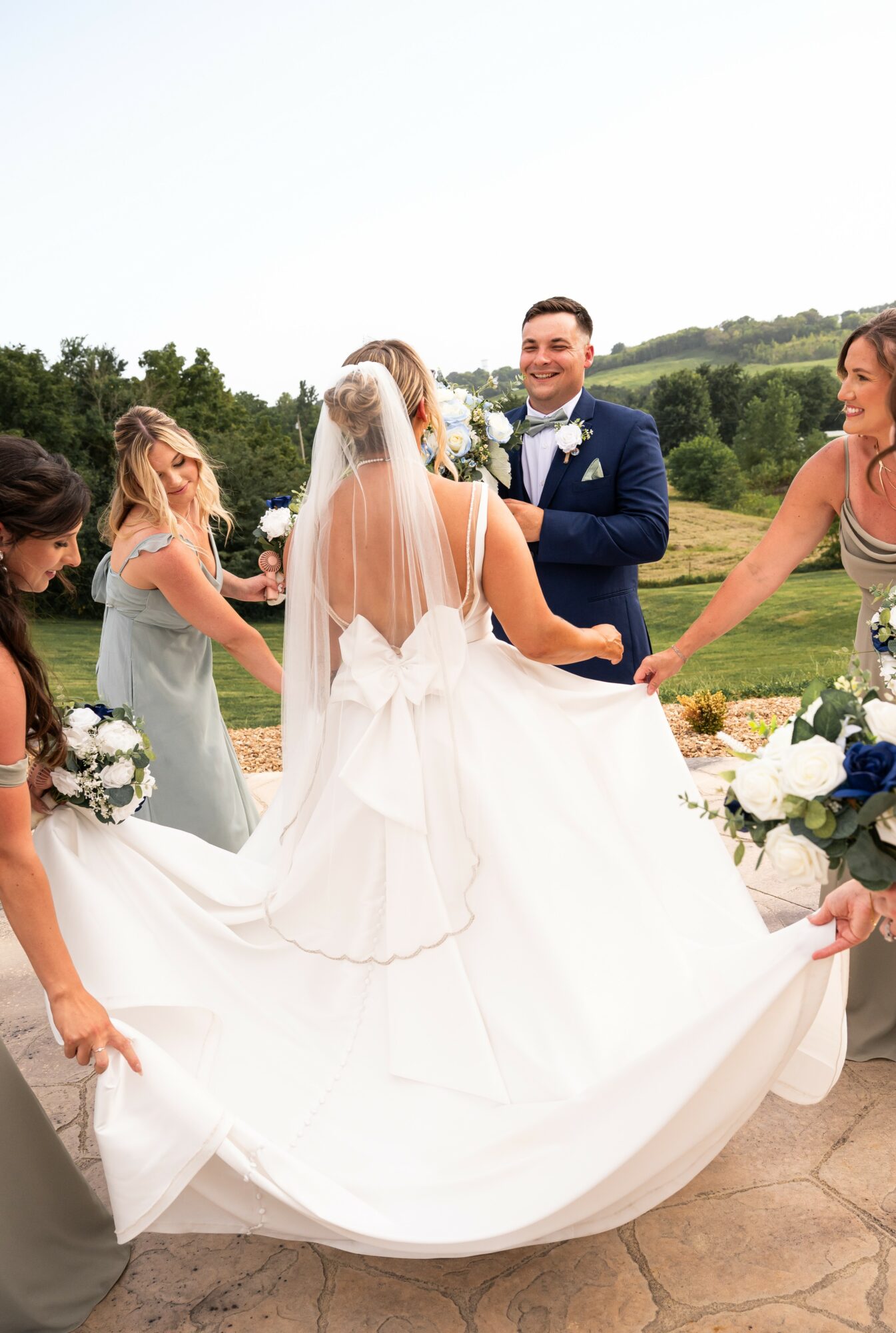 Bride in white dress with veil holding hands with groom in blue suit, surrounded by bridesmaids outdoors.