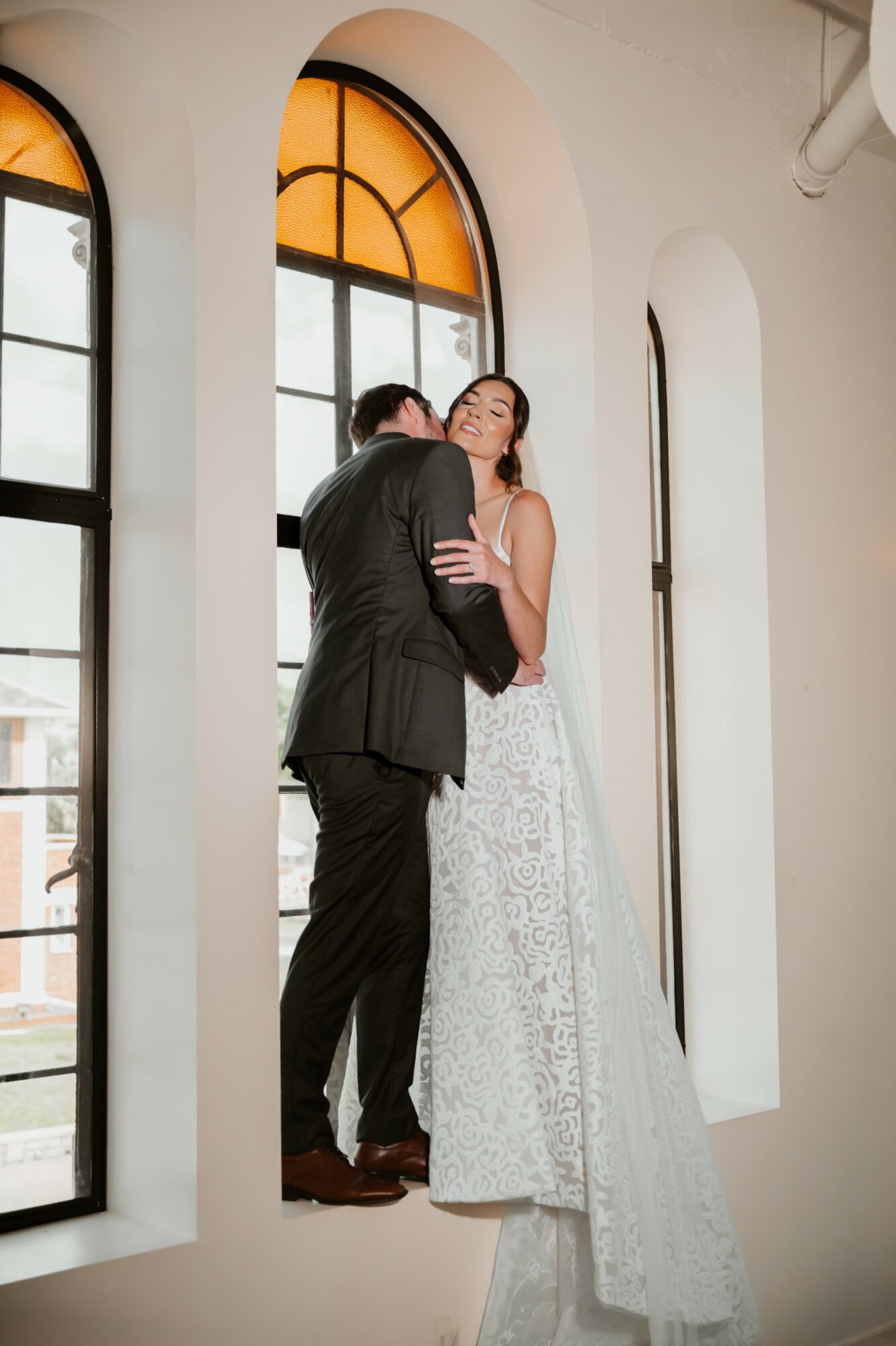 A couple dancing inside a bright room with large arched windows, one in a suit and the other in a white dress.