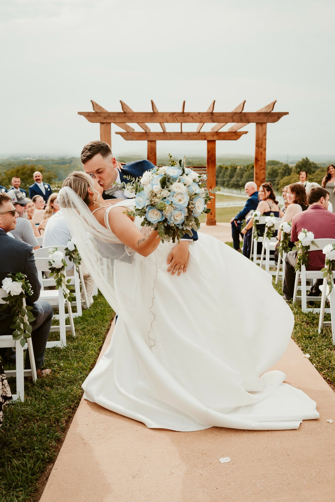Bride and groom kissing outdoors during wedding ceremony, guests seated on chairs, wooden arch in background, cloudy sky.