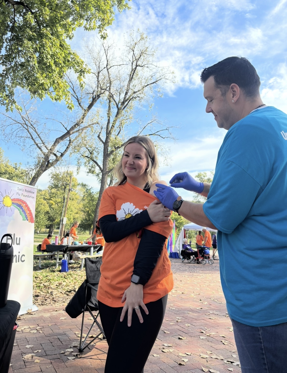 A woman in an orange shirt smiling as a man in a blue shirt and gloves interacts with her outdoors.