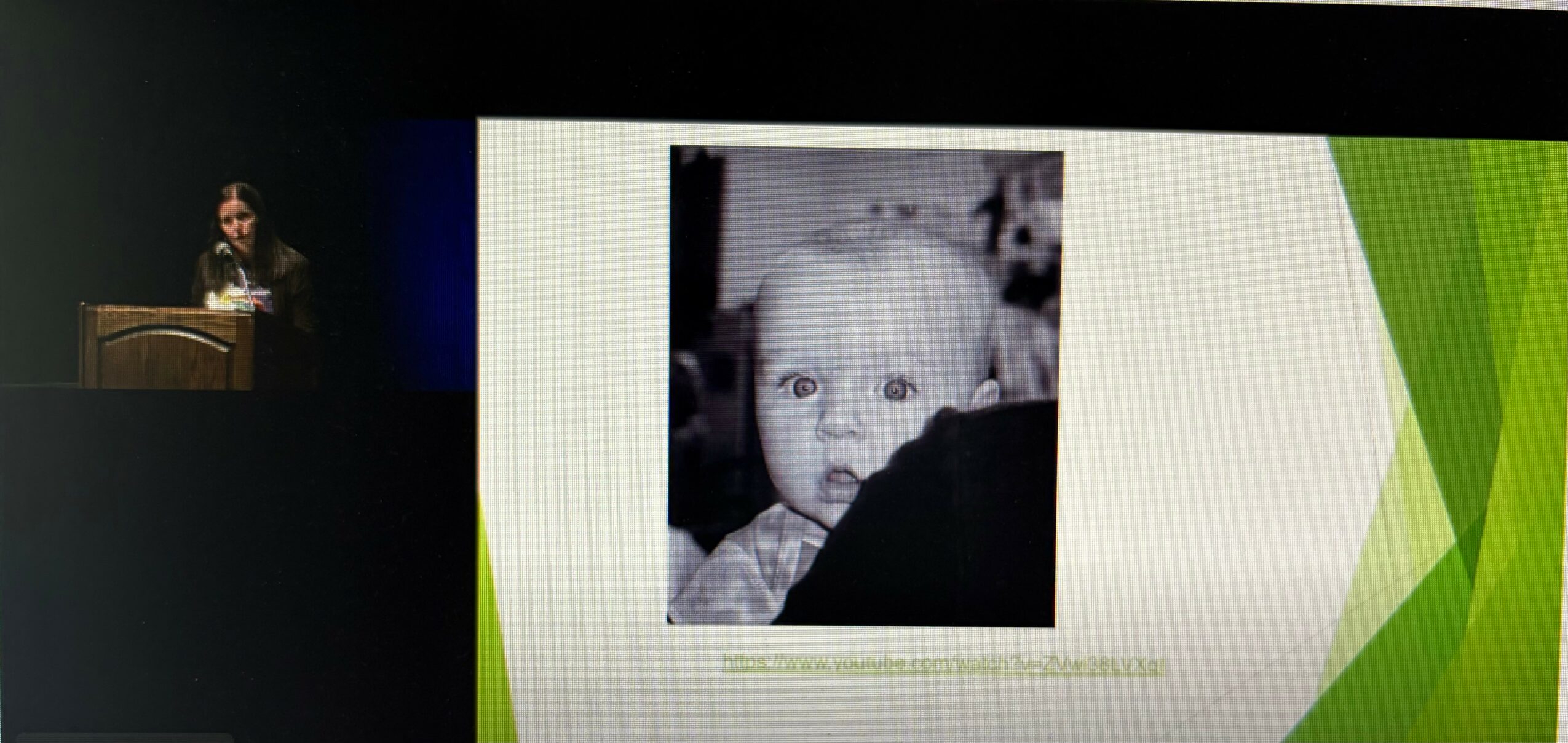 A woman presents at a podium with a large screen displaying a black-and-white photo of a young child with wide eyes.