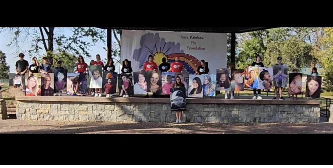 Group of children and adults on stage with photos displayed behind them, outdoors with trees and a stone wall.