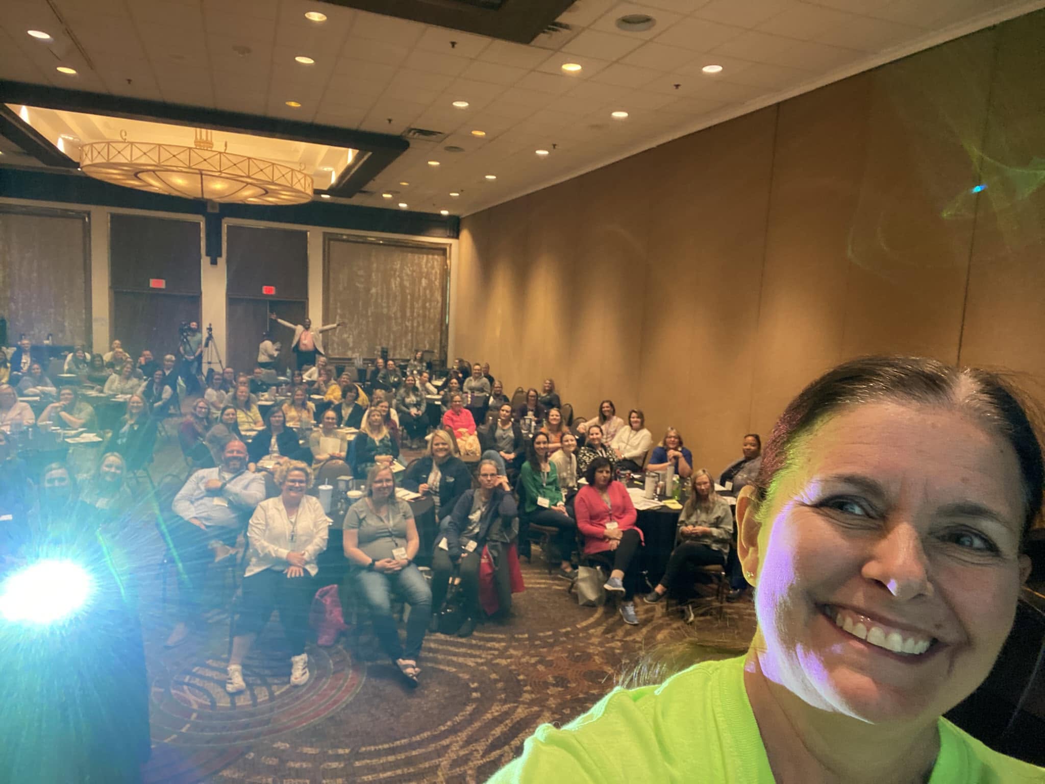 Smiling woman in bright yellow shirt taking a selfie at a large conference or event with many seated attendees in a ballroom.