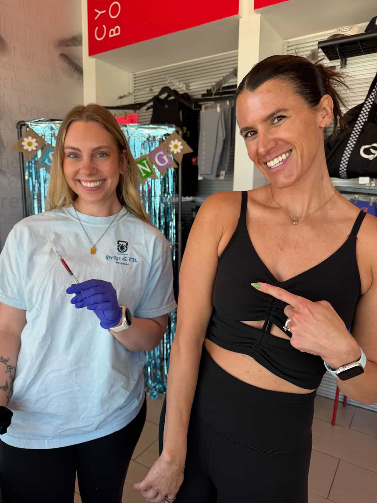 Two women smiling indoors, one wearing a light blue shirt and gloves, the other in a black top, pointing at each other.