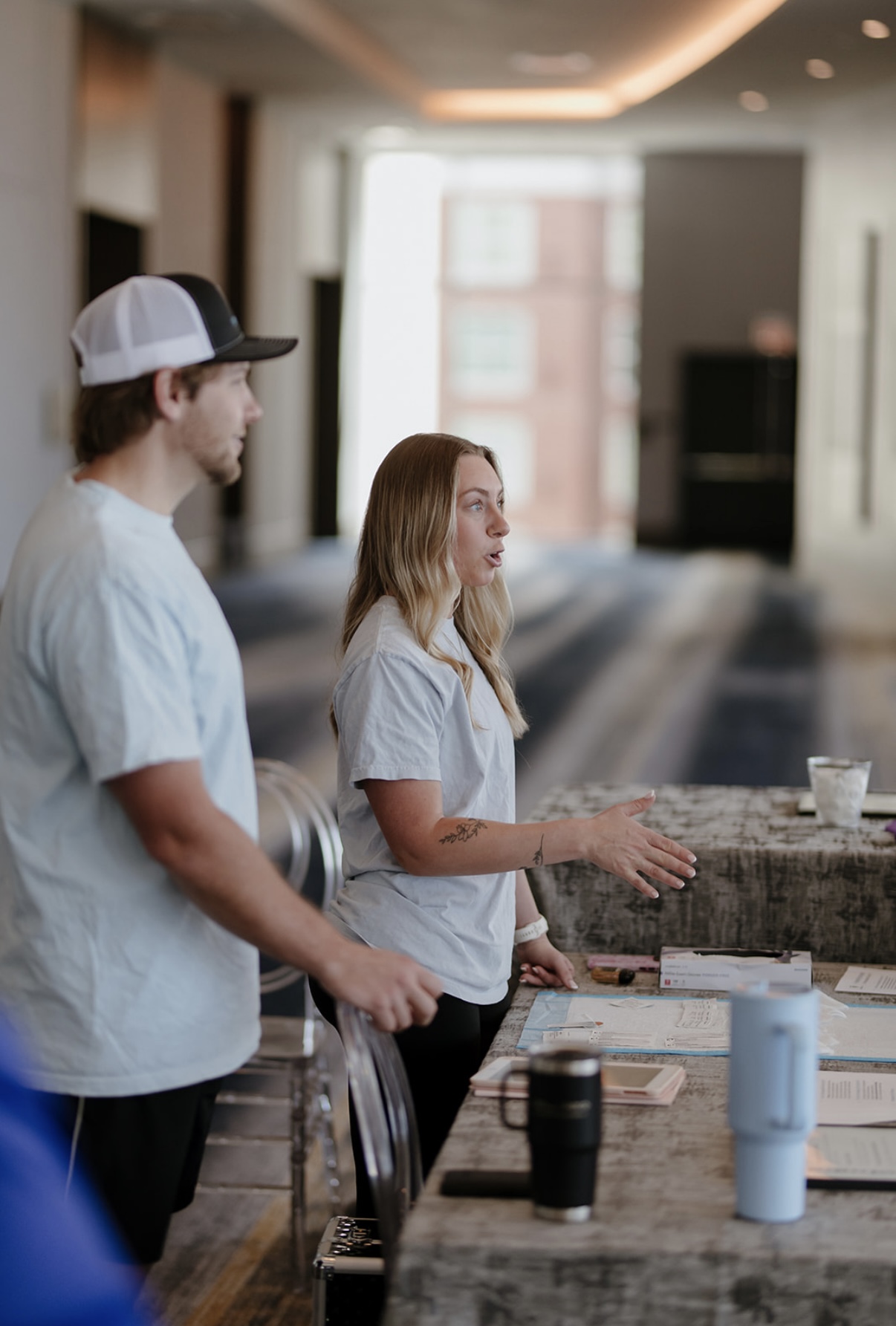 Two people standing at a counter in a modern indoor space, with a window and blurred background behind them.