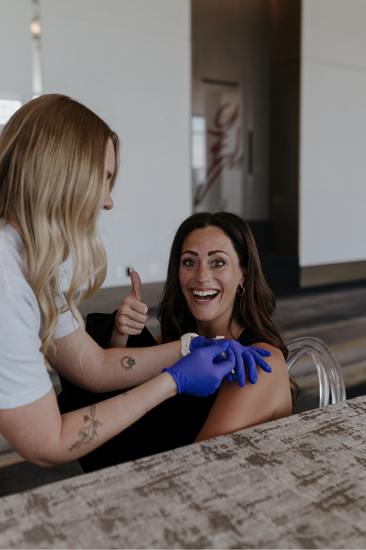 A woman smiling and giving a thumbs-up while a person with blonde hair and blue gloves works on her arm.