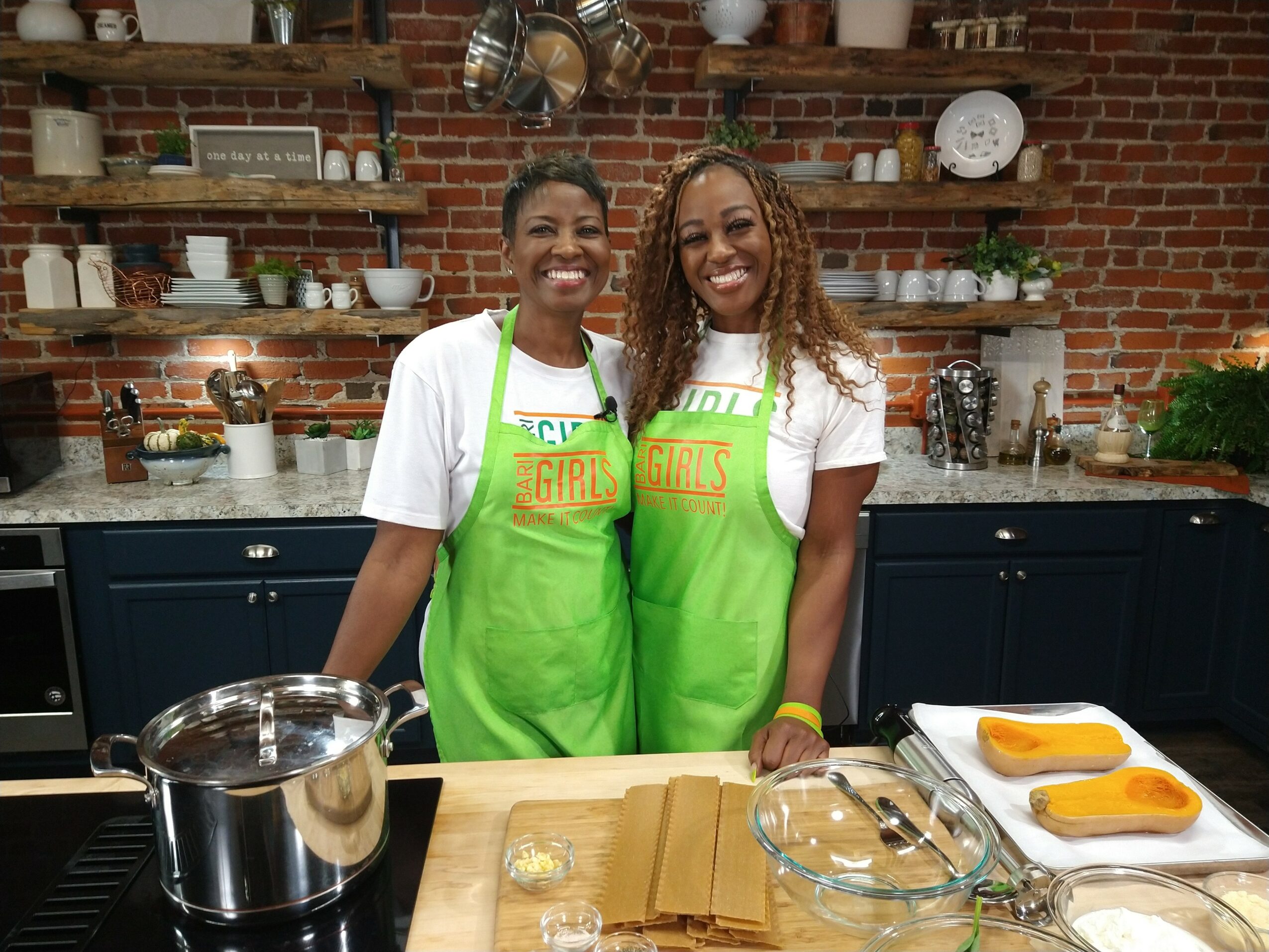Two women wearing green aprons stand in a kitchen with brick walls, smiling, with cooking utensils and ingredients on the counter.