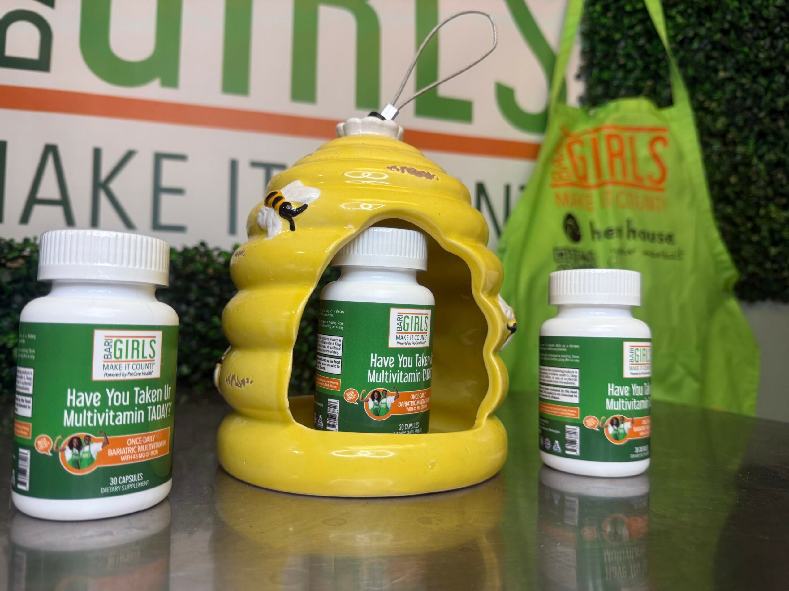 Three bottles of multivitamins and a yellow bee-shaped container on a table, with a green bag and a sign in the background.