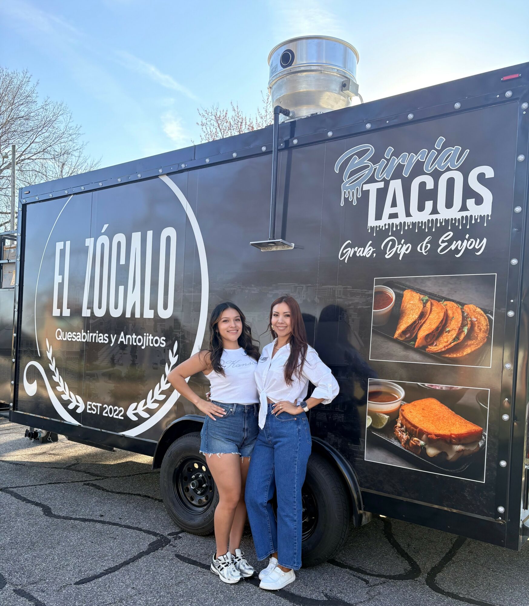 Two women stand in front of a black taco food truck, smiling, with clear sky and trees in background.