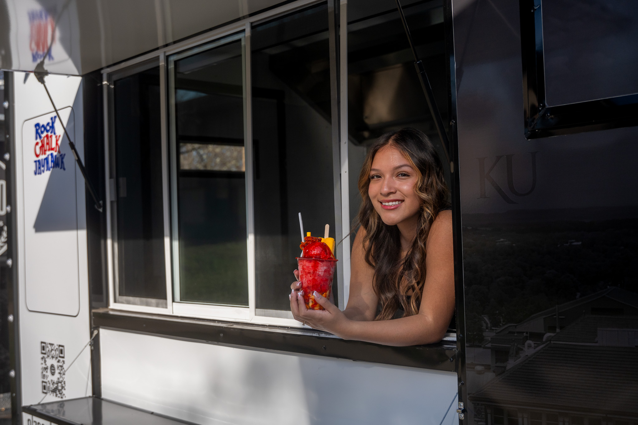 Young woman with long hair smiling and holding a red drink with fruit and a straw, looking out of a window.