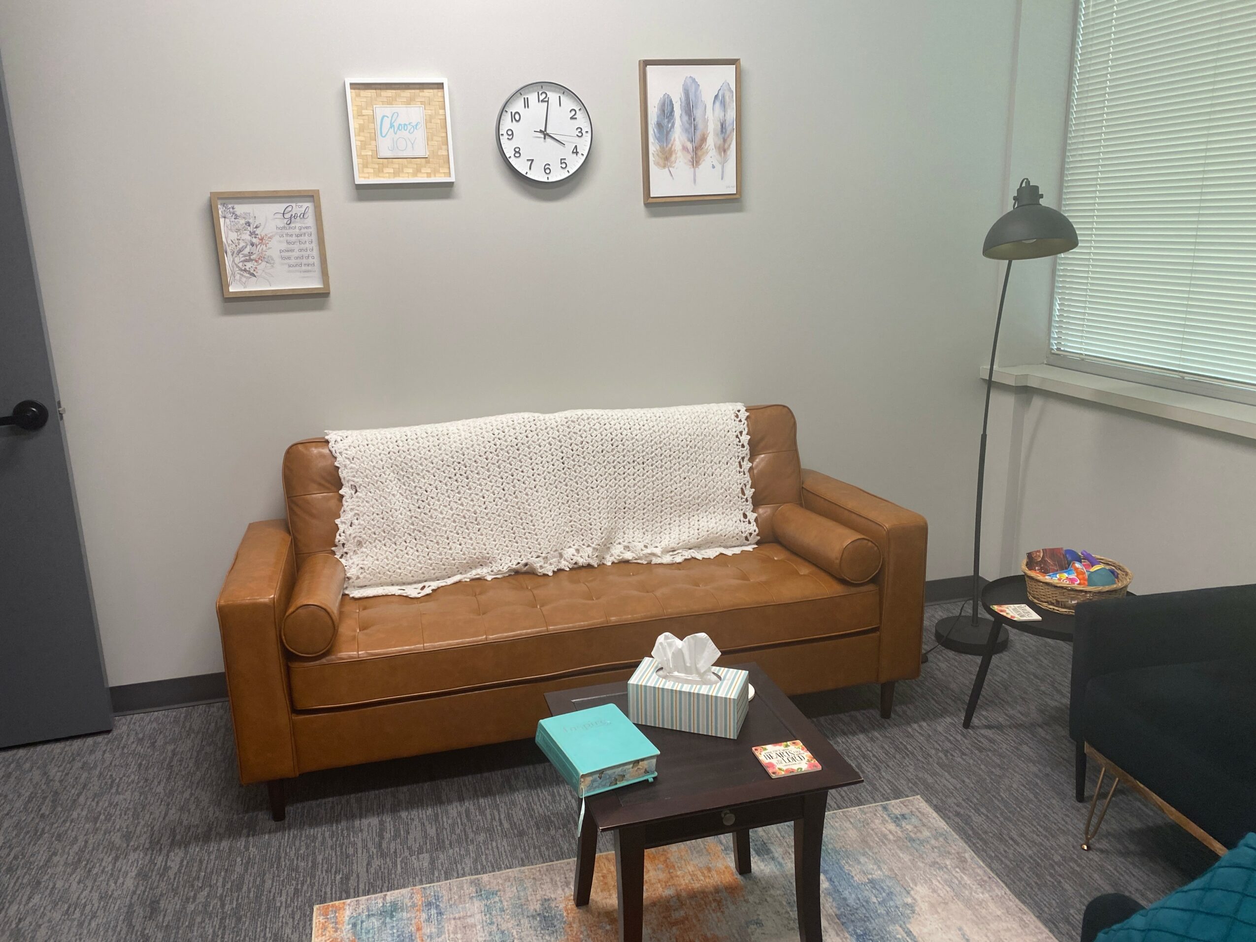 Living room with a brown sofa, wall art, clock, window, and a floor lamp, with a small table and chair nearby.