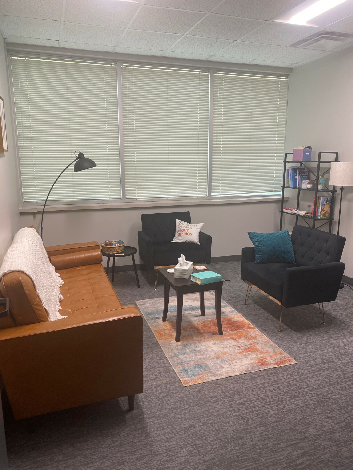Living room with black sofa, small wooden table, colorful rug, and bookshelf near window with blinds.