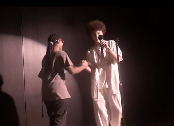 Two people on stage, one singing into a microphone, the other holding their hand, with a dark background.