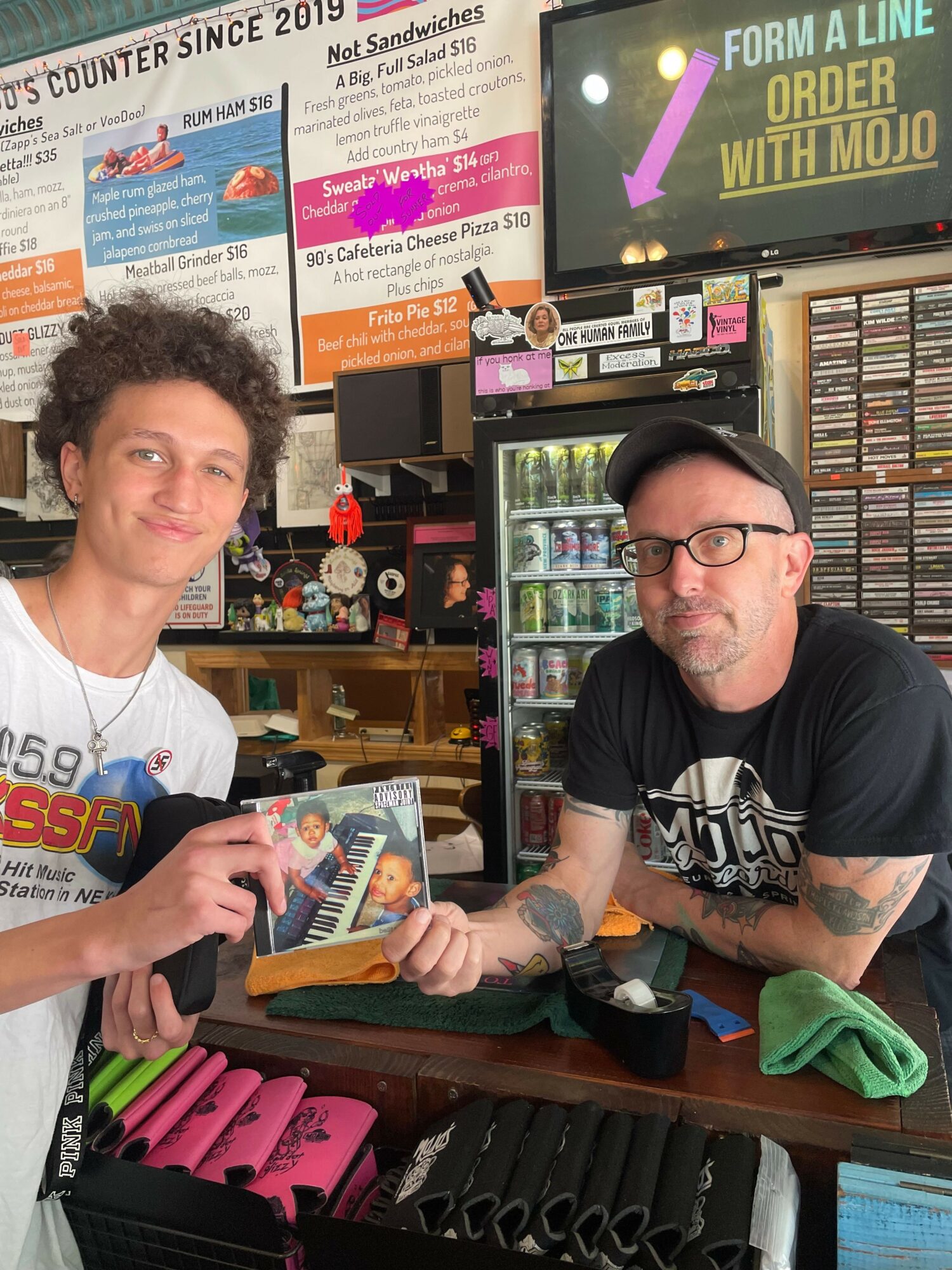 Two people at a store counter, one holding a book, shelves with DVDs and posters in the background.