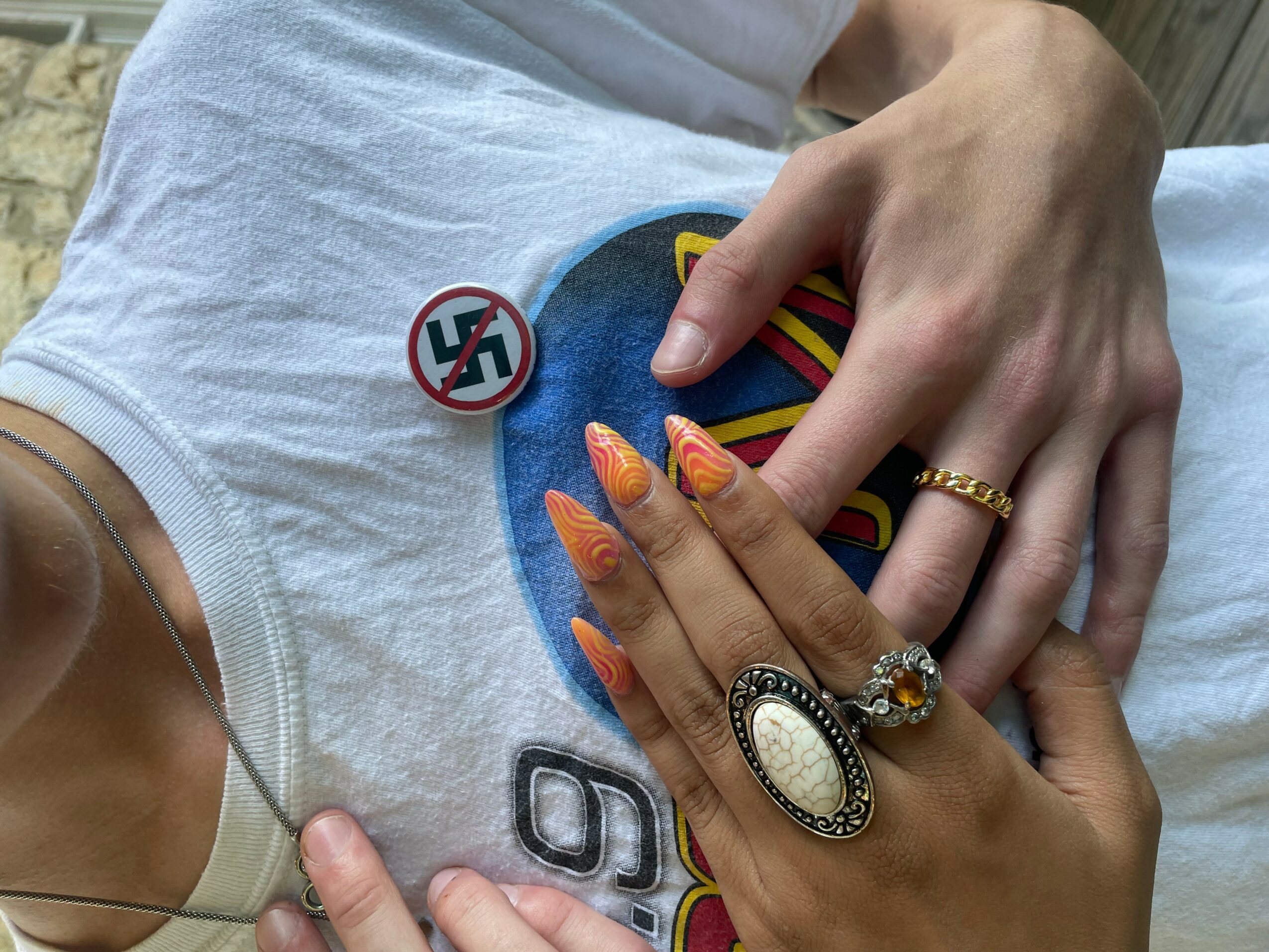 Person wearing a white shirt with Superman logo, multiple rings, and long decorated nails, with a hand resting on chest.