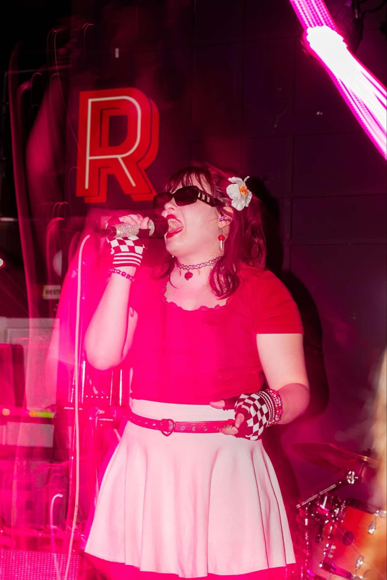 Woman in vintage style outfit with glasses, gloves, and a flower in her hair, holding a microphone on stage with neon lights.