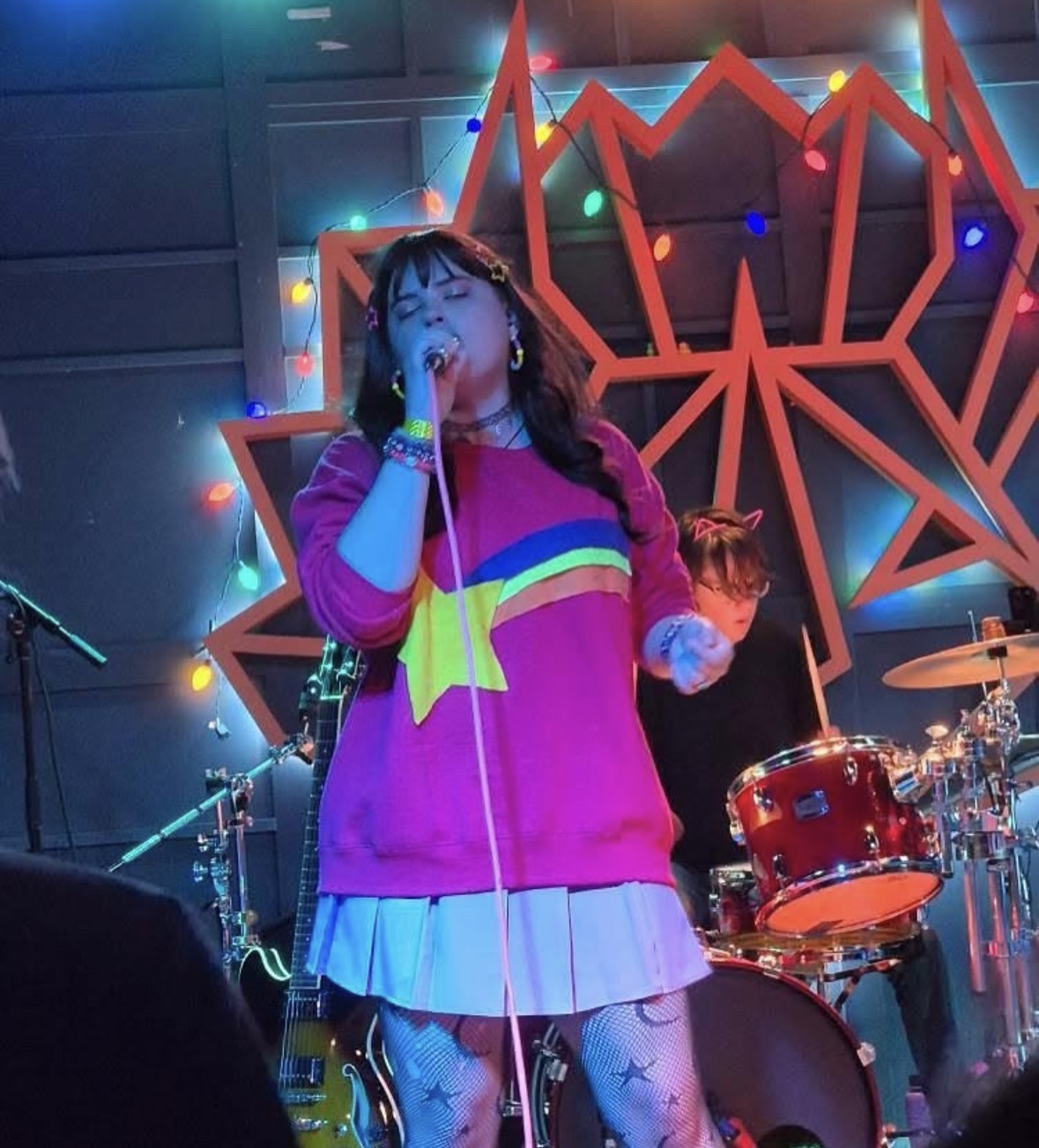 Performer singing on stage with colorful lights and a large geometric backdrop, band members playing instruments in the background.