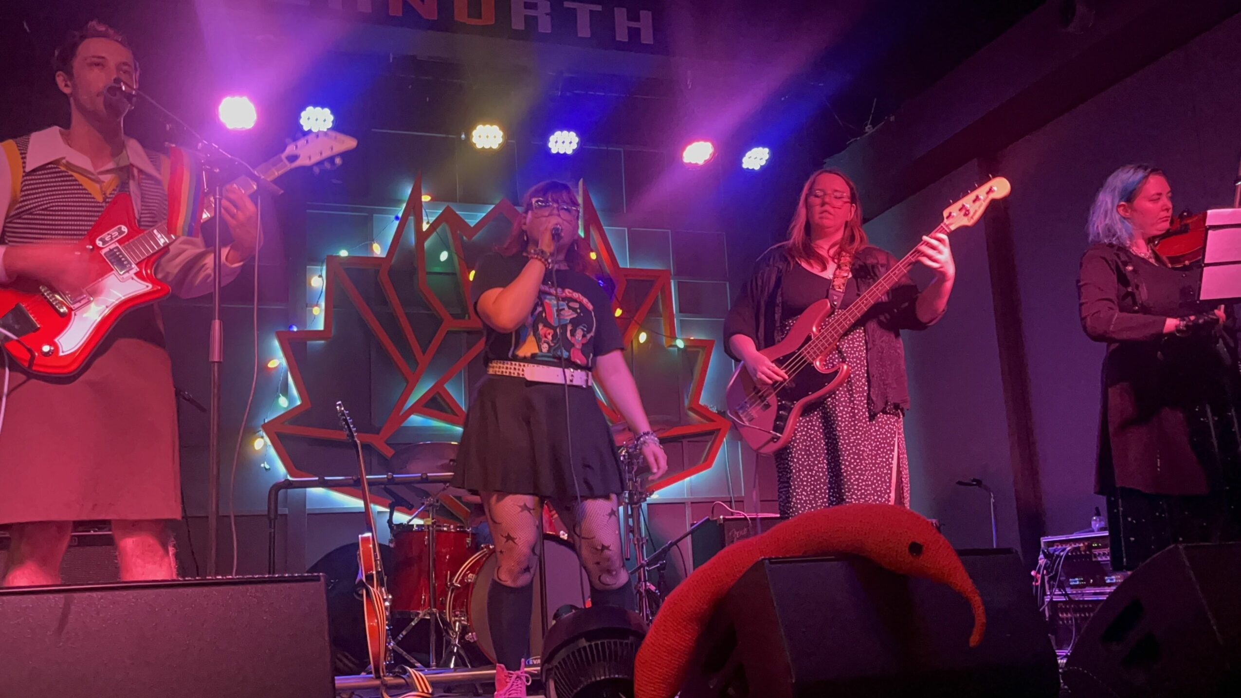 Four musicians perform on stage with colorful lights and a geometric backdrop, playing guitars and singing.