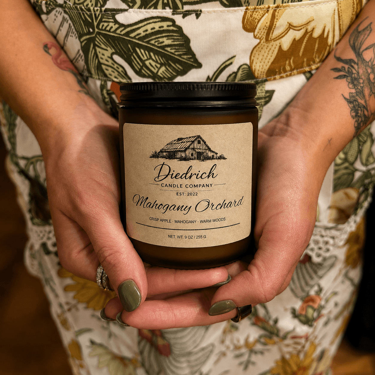 Person holding a candle jar with a label reading 'Diedrich Candle Company Mahogany Orchard' in front of floral-patterned clothing.