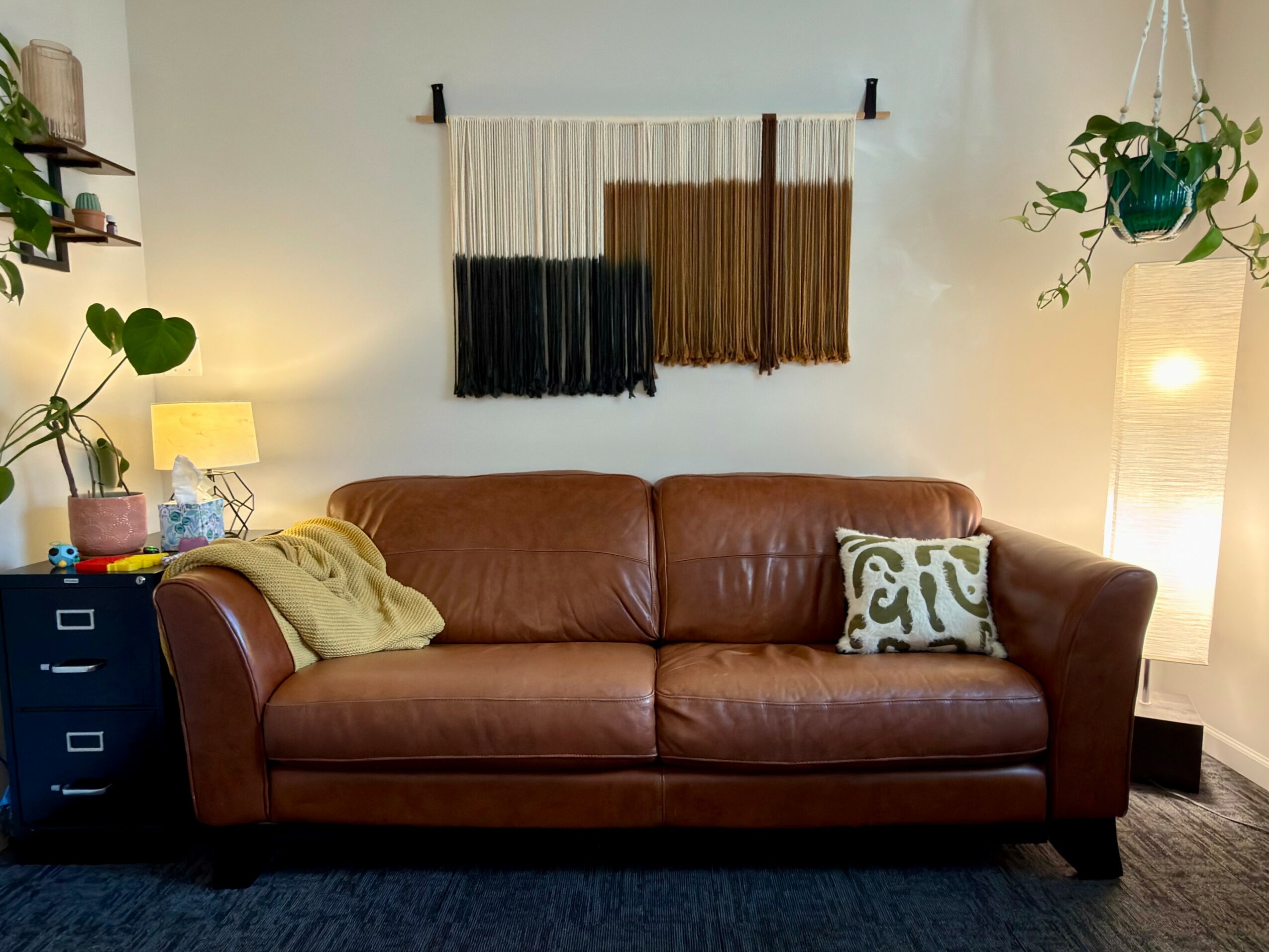 Living room with a brown leather sofa, wall hanging, plants, and a lamp, illuminated by warm lighting.