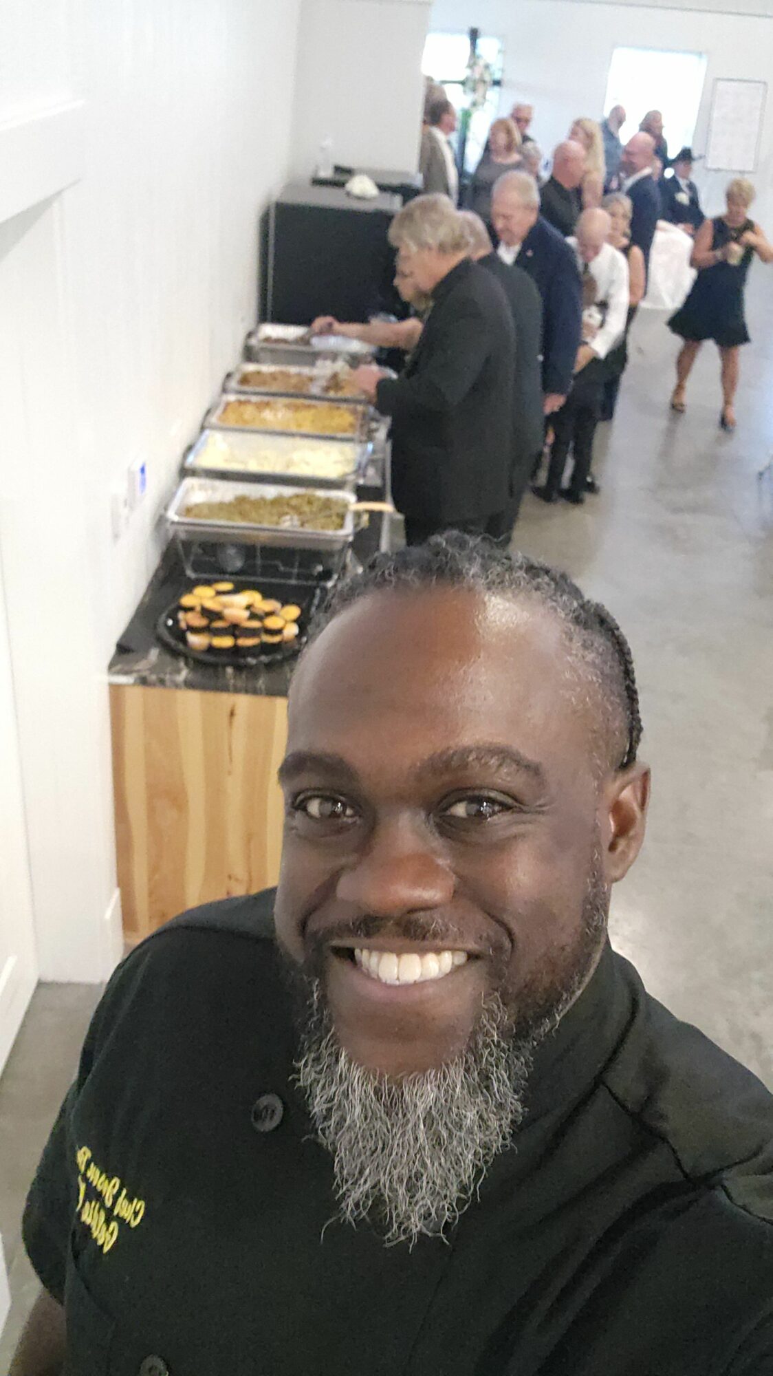 Smiling man with a beard in a black shirt, standing in front of a buffet table with food, in a busy room.