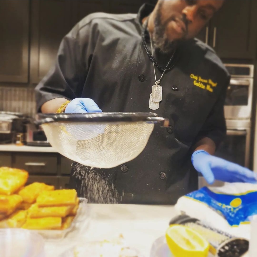 Chef sifting flour into a bowl, wearing a black uniform and blue gloves, in a kitchen setting with food in foreground.