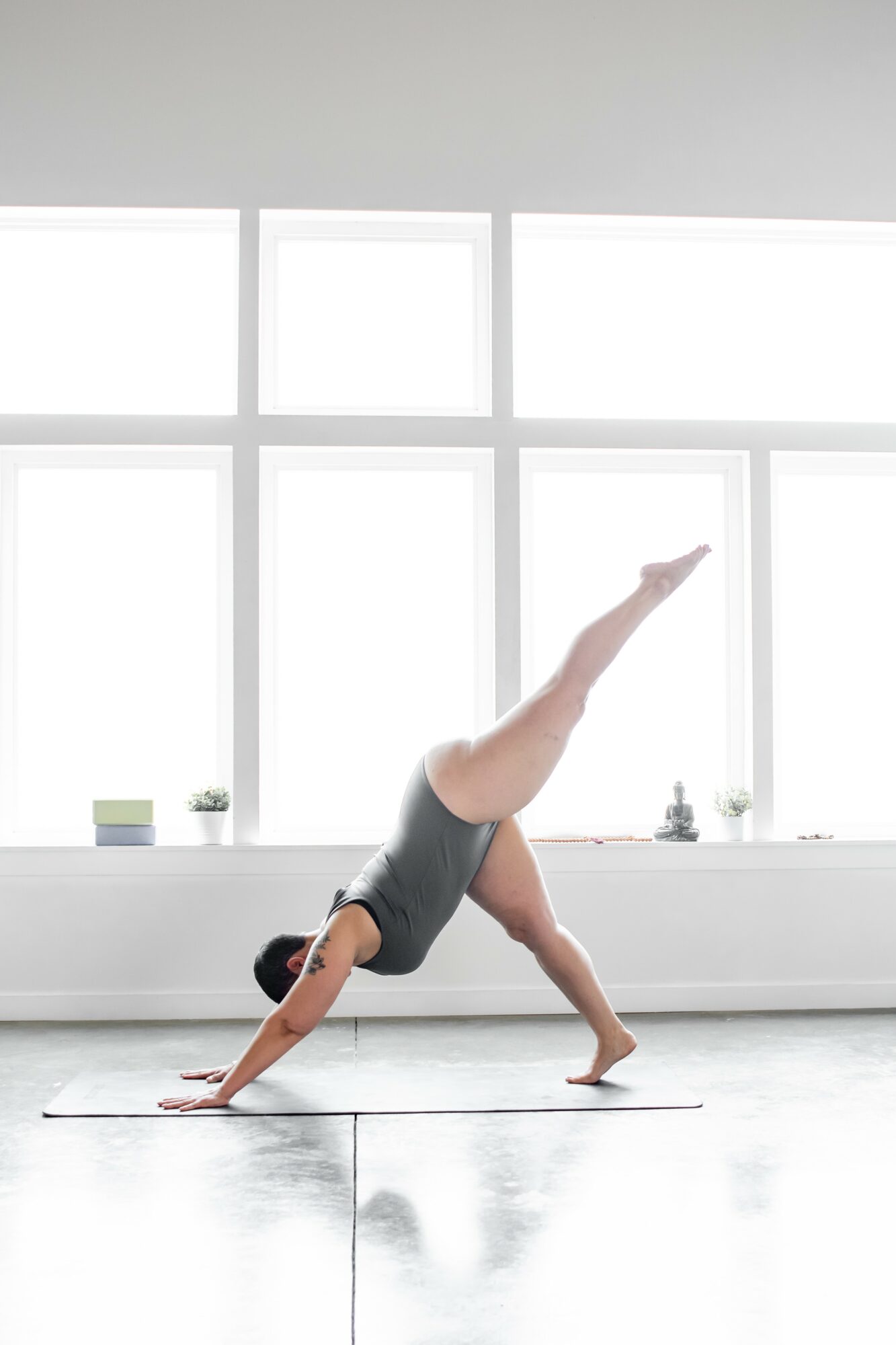 Person practicing yoga in a bright room with large windows, performing a downward dog pose with one leg raised.