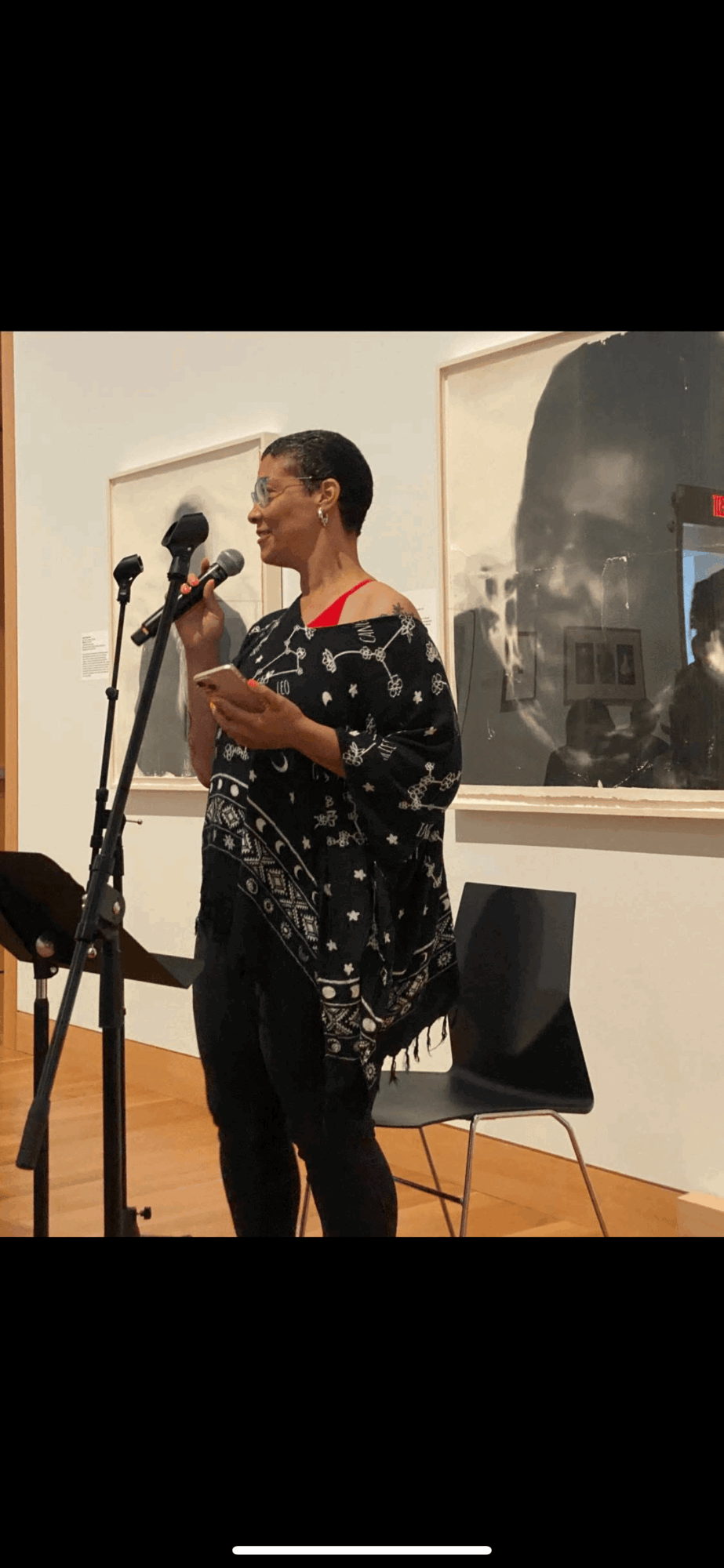 Person speaking into microphone, holding notes, in an art gallery with framed artwork on the wall behind.