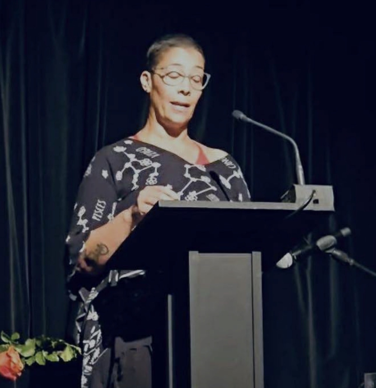 Woman with glasses speaking at a podium with a dark curtain background.