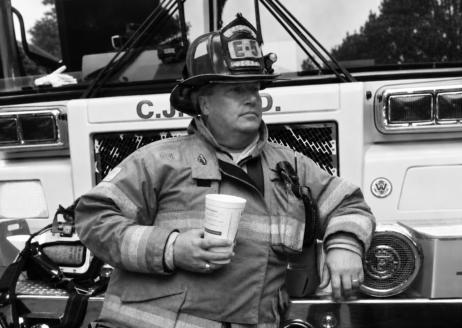 Firefighter in uniform holding a cup, standing in front of a fire truck, looking to the side.