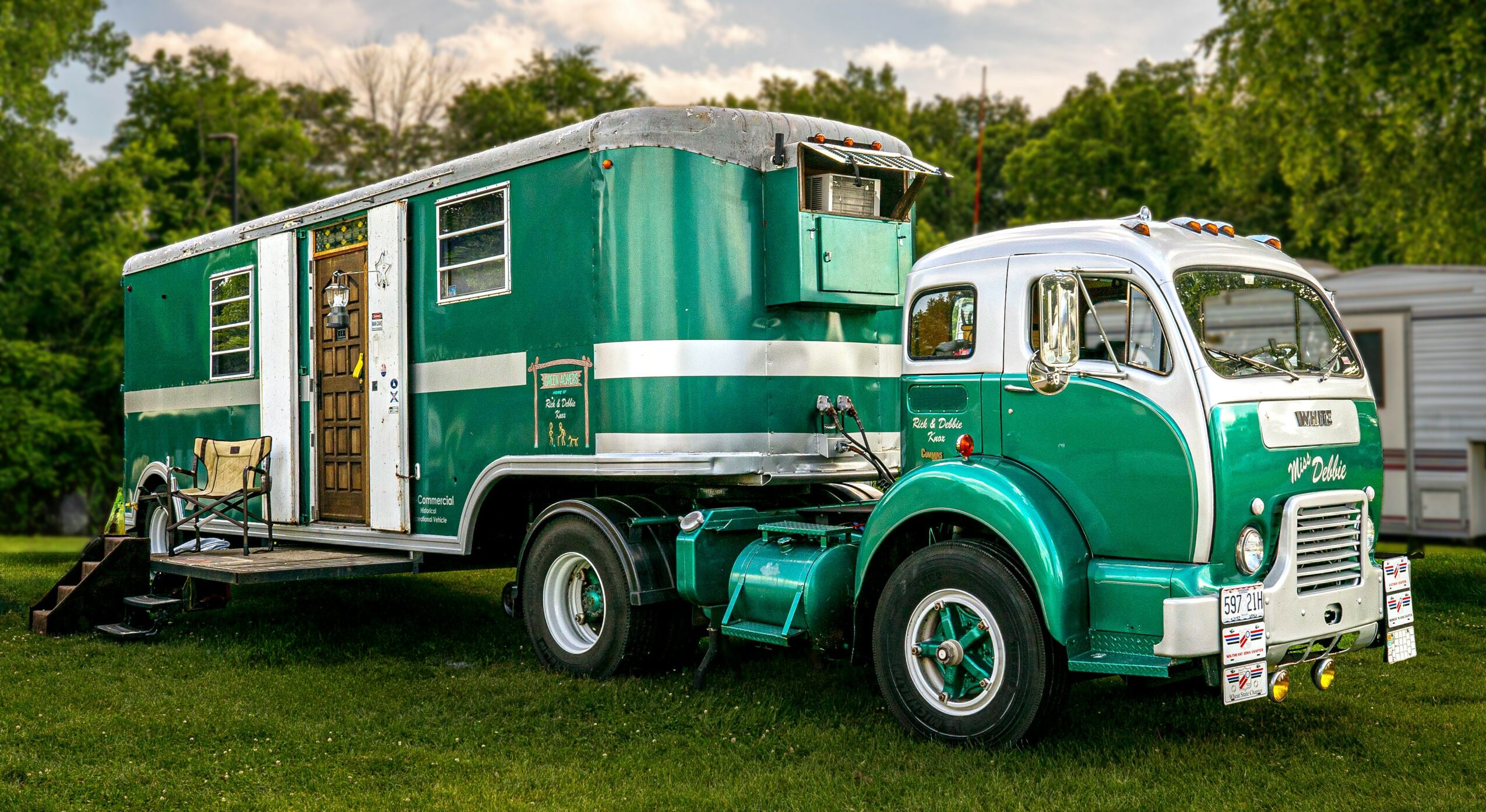 Green and white vintage truck with a large attached trailer parked on grass, trees in background.