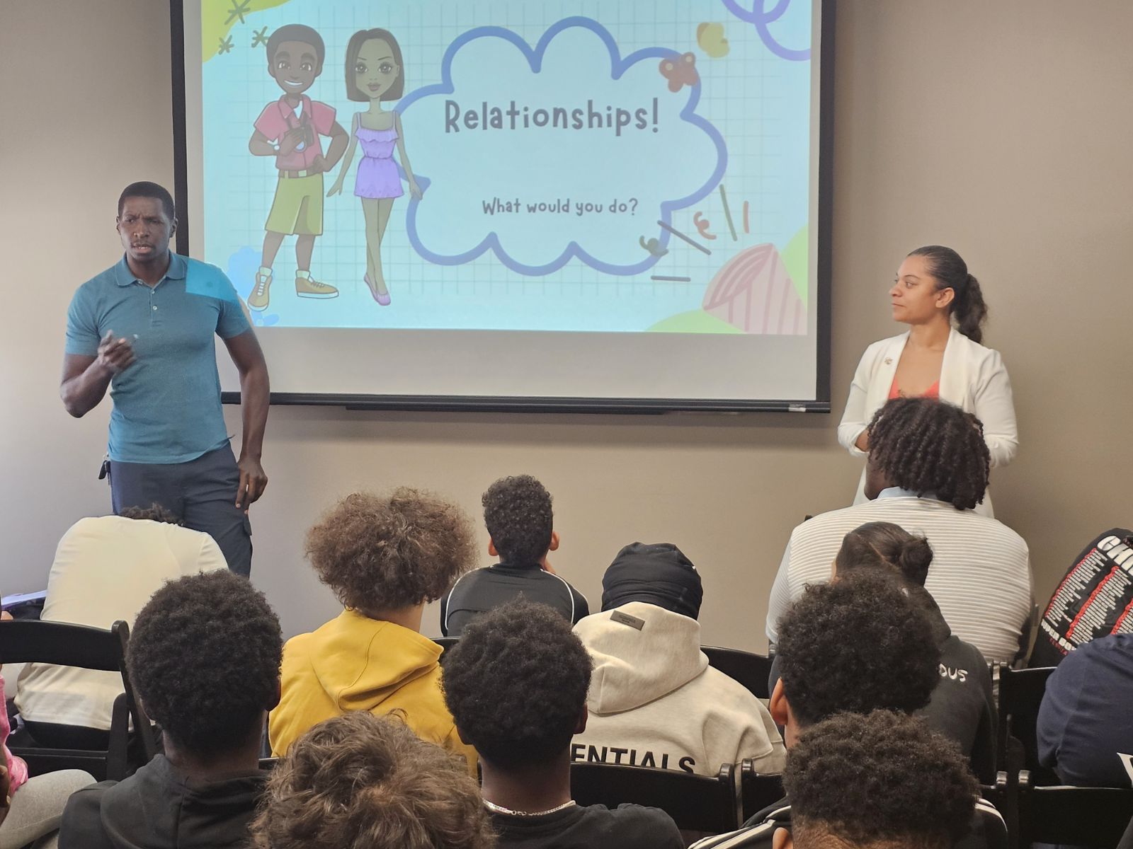 Two presenters stand in front of a screen with a slide about relationships, audience seated and listening attentively.