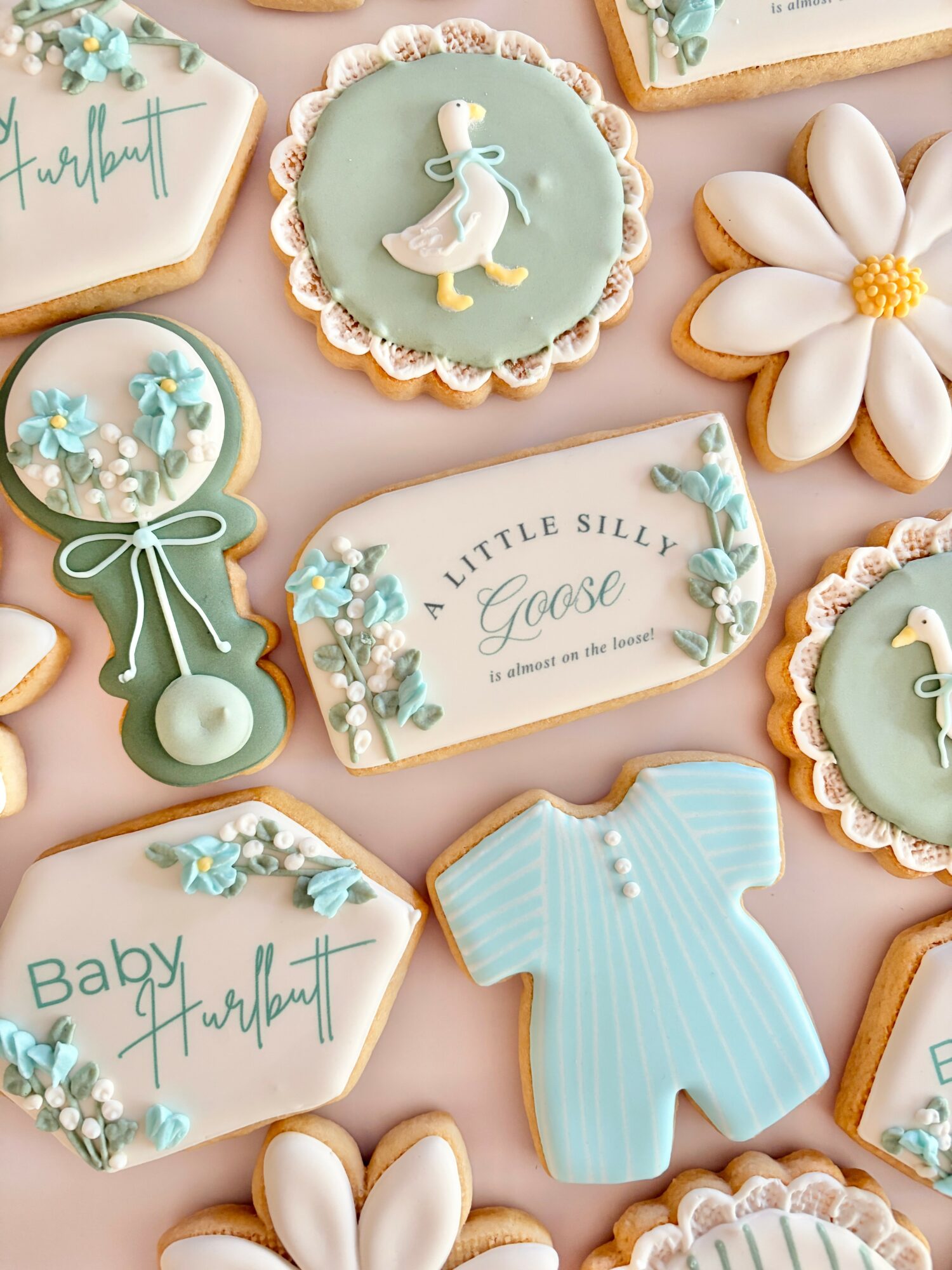Decorative cookies with baby and nature themes, including a duck, flowers, a onesie, and text, on a pink background.