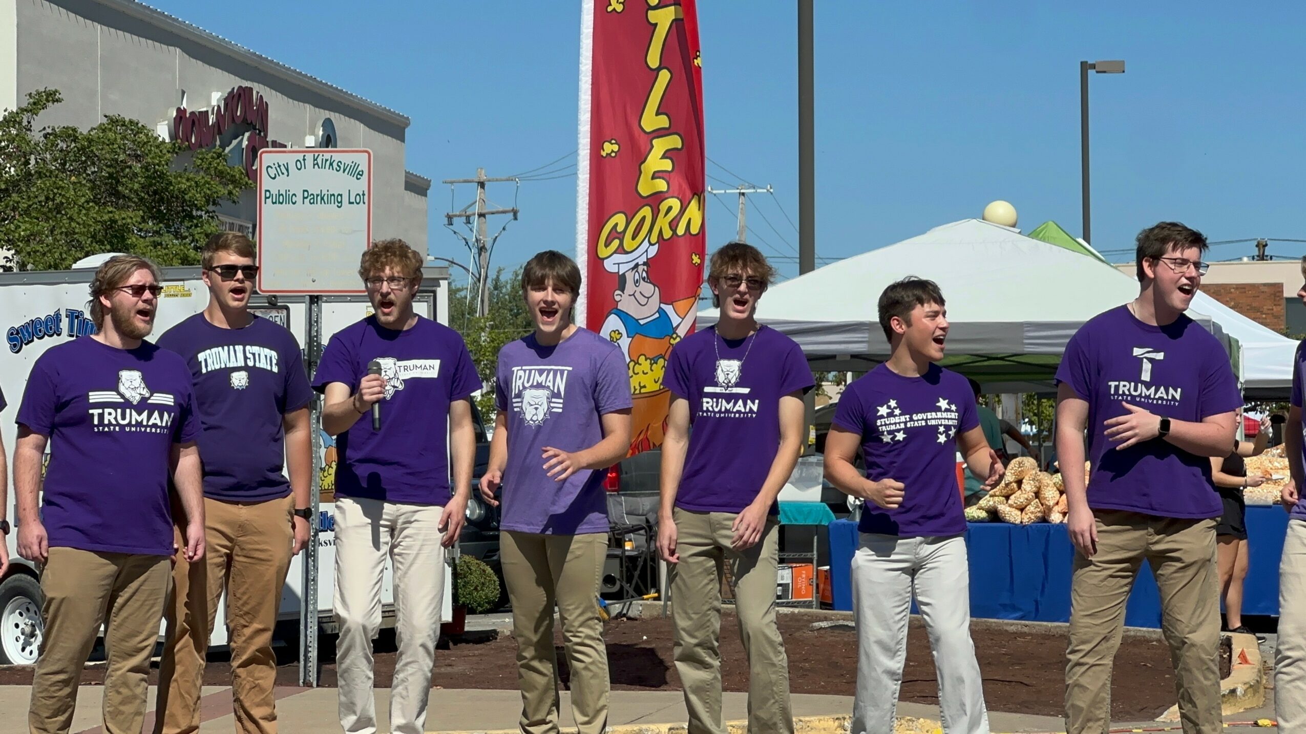 Seven people wearing purple shirts stand outdoors in front of a tent and a flag with a yellow and red sign, under a blue sky.
