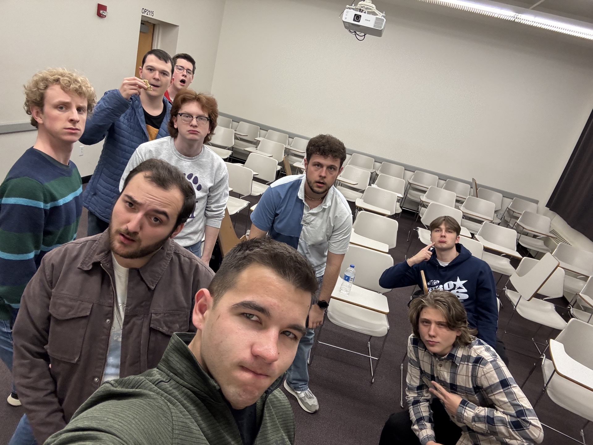 Group of eight young people in a classroom with empty desks and chairs, some looking at the camera, one making a peace sign.