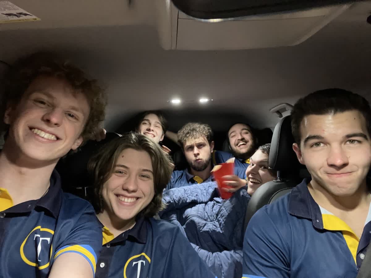 Group of seven young people smiling and sitting in a car, some holding red cups, wearing matching blue and yellow shirts.