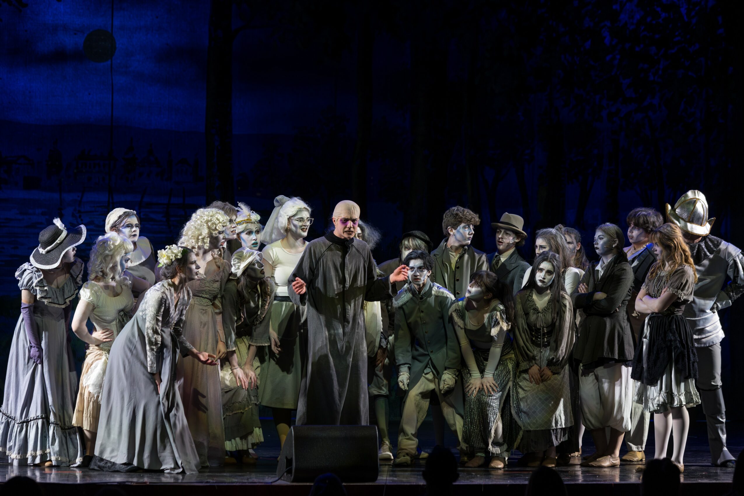 Group of people on stage, some in costumes, in a theatrical performance setting, with dark background and stage lighting.