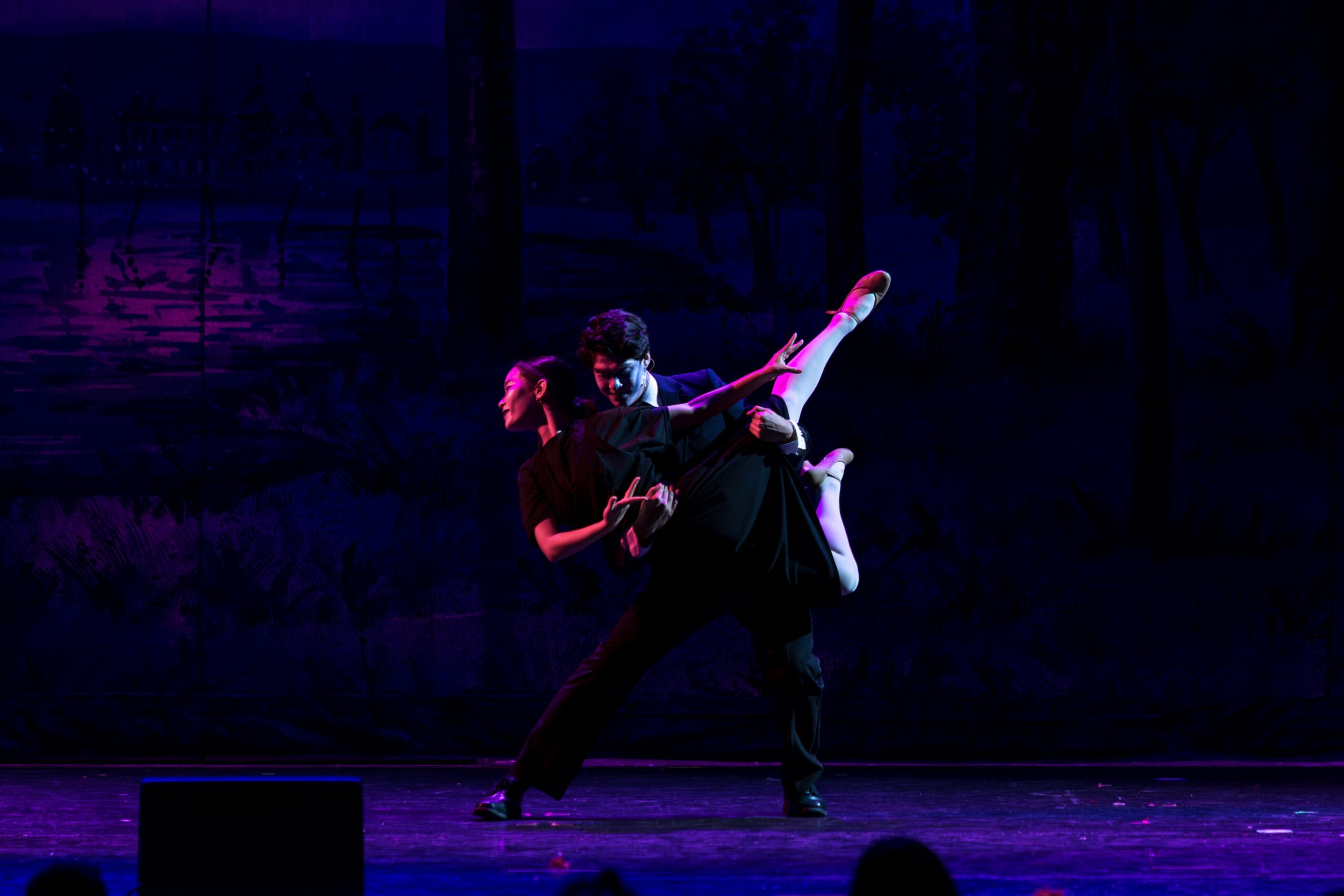 Two ballet dancers perform on stage, one lifting the other in an extended pose, with dark background lighting.
