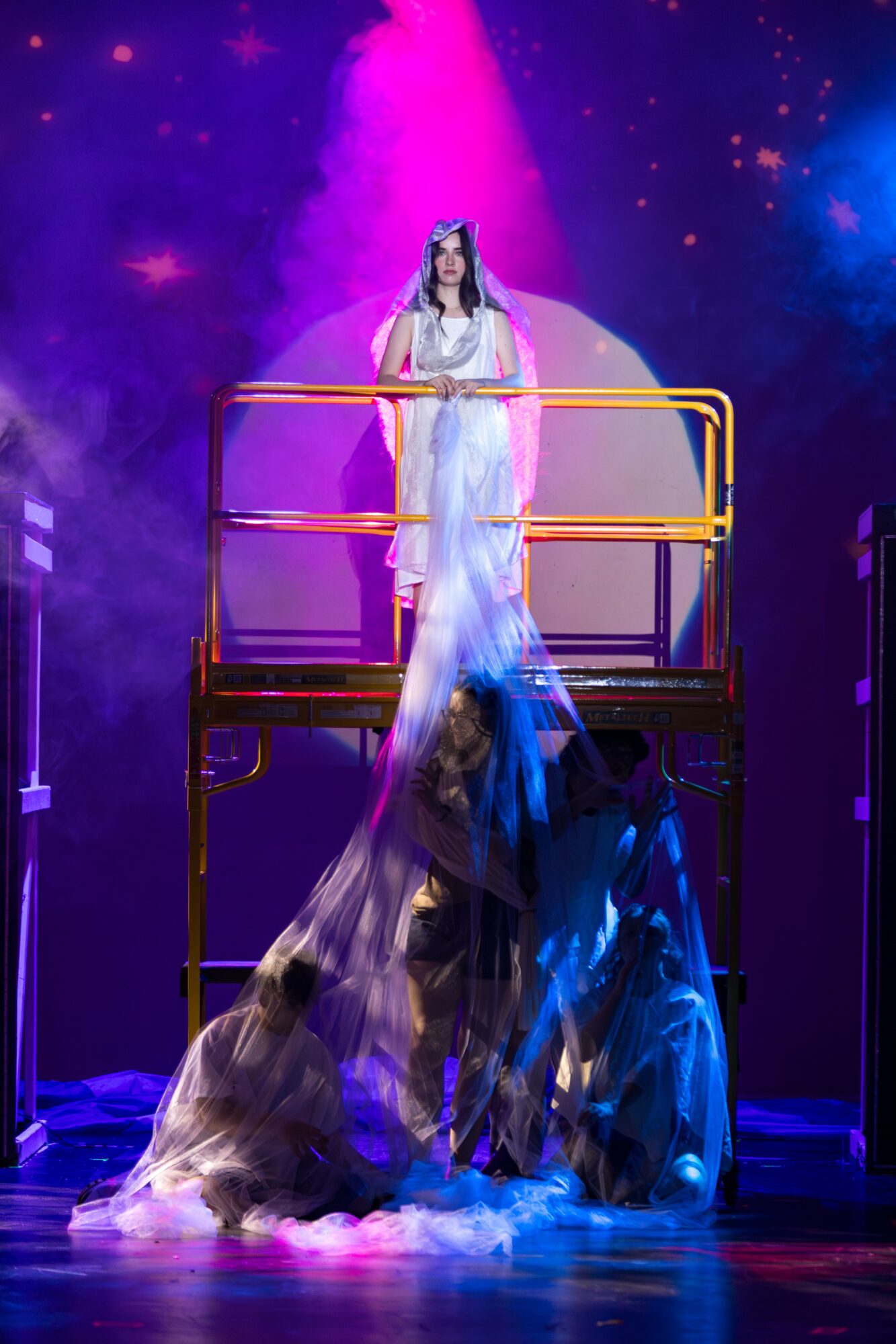 Performer in white dress stands on a platform with colorful lights and flowing fabric below.
