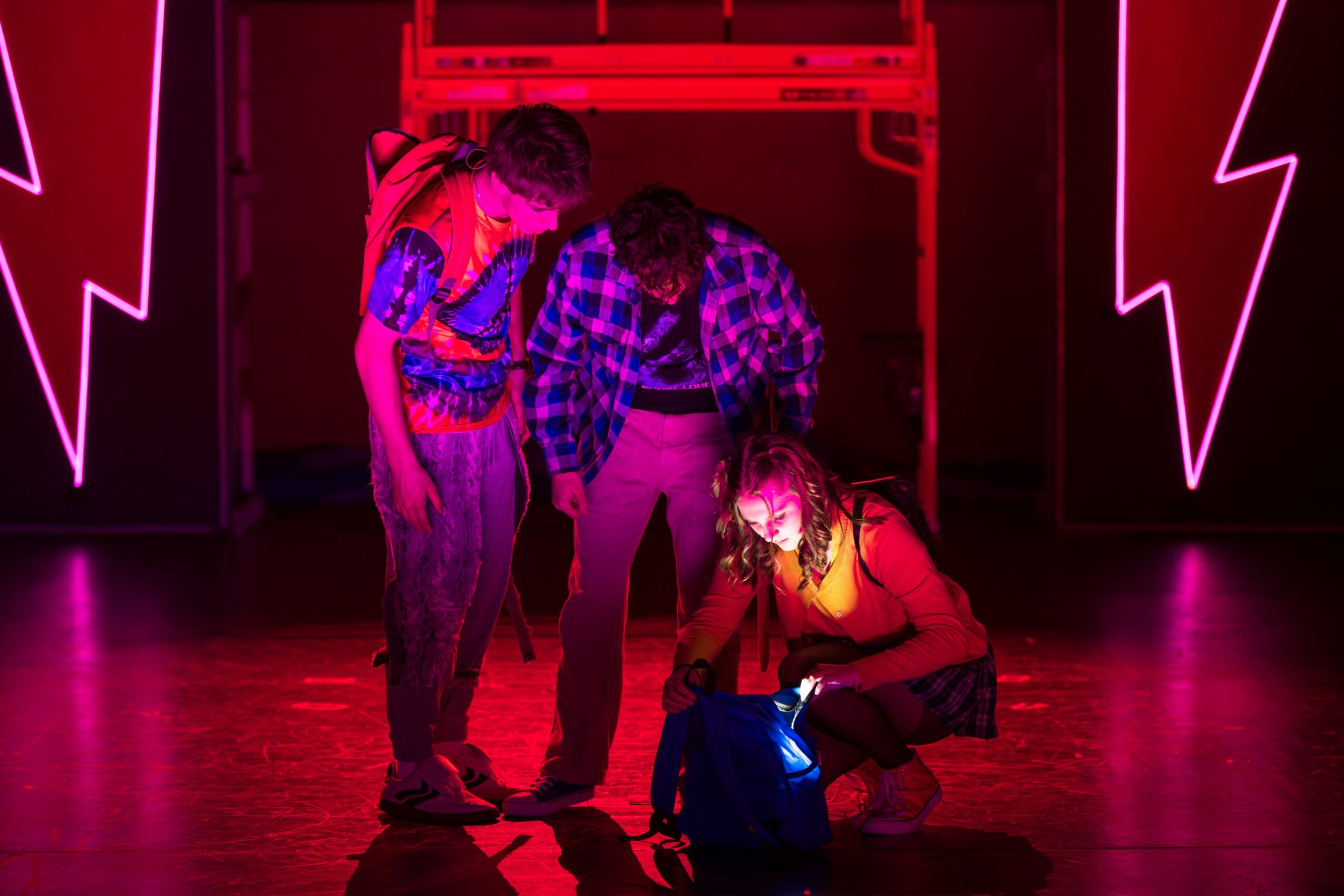 Three people in a dimly lit room with neon lightning bolt decorations, one crouching and two standing, all looking at something on the floor.