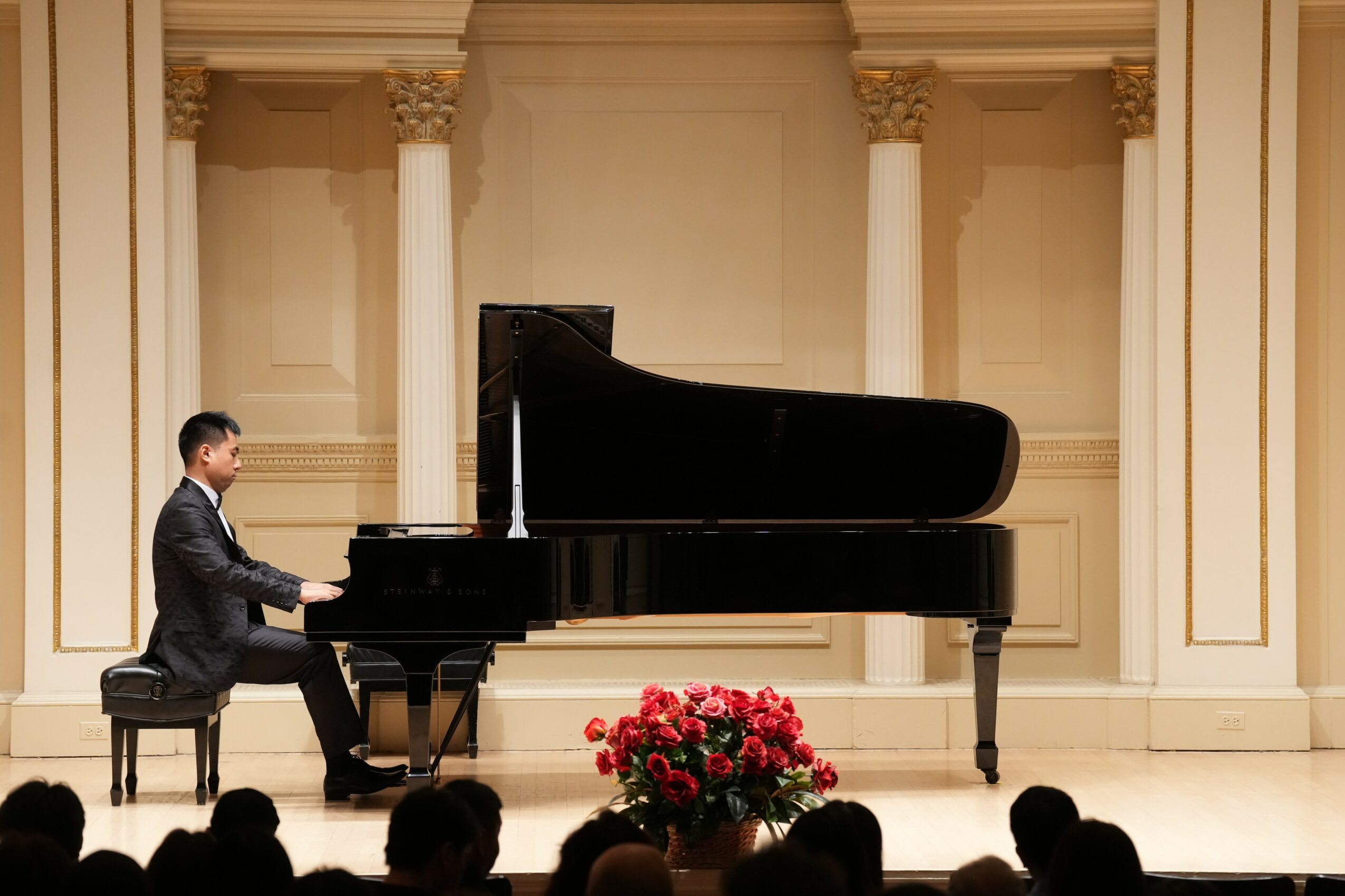 A person playing a grand piano on stage with columns and a floral arrangement in front.
