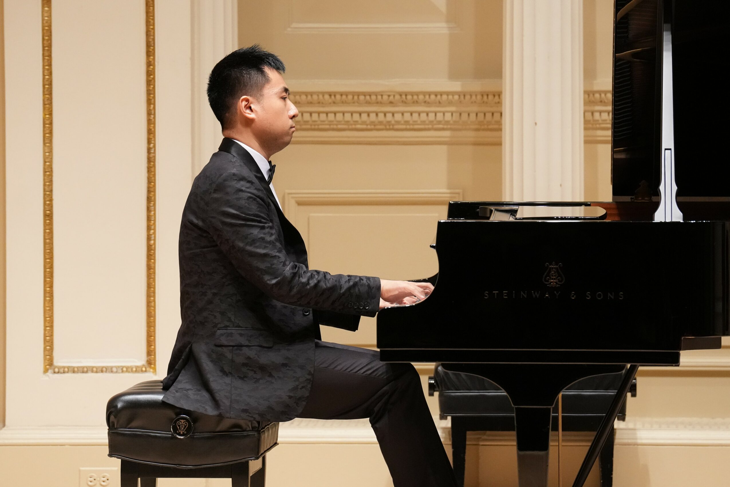 Young man playing piano in a formal room with ornate decor, seated on a bench, facing right.