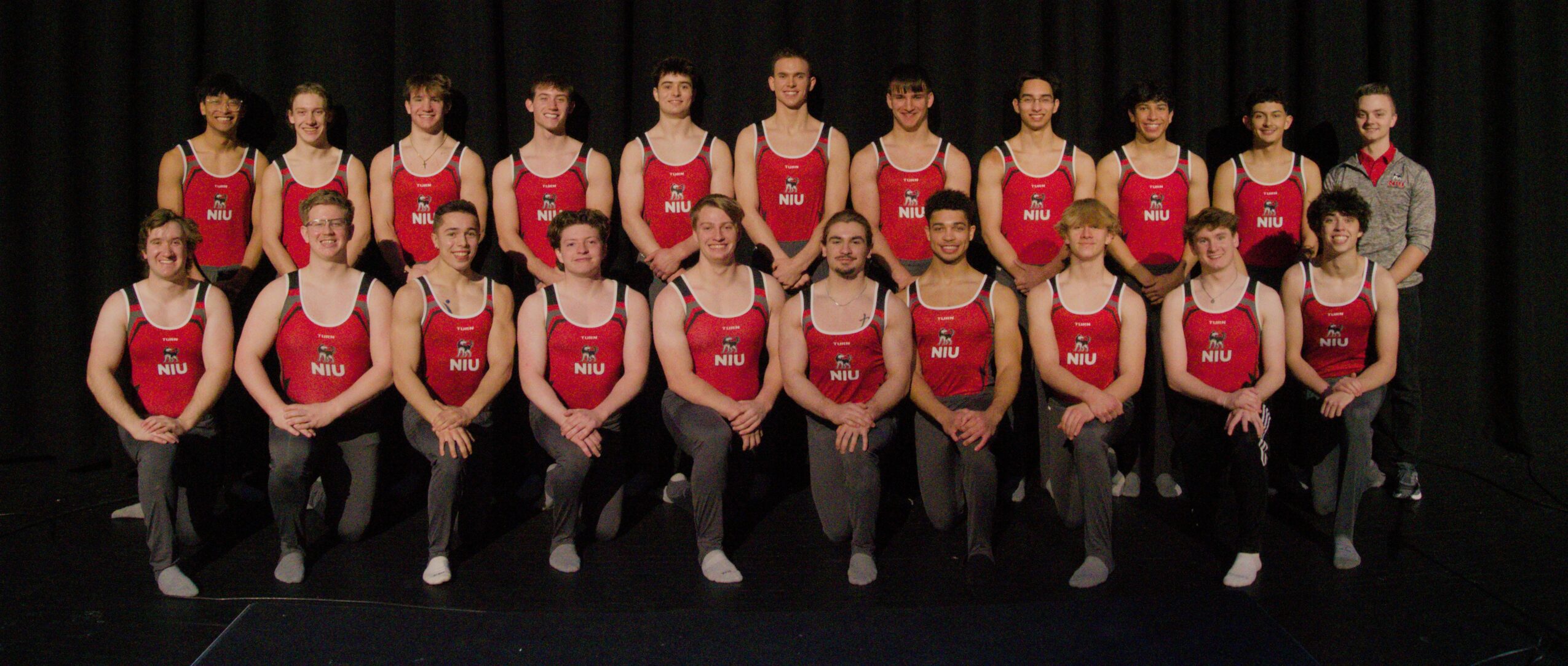 Group of 20 women in red and black athletic outfits posing on stage with a black curtain background.