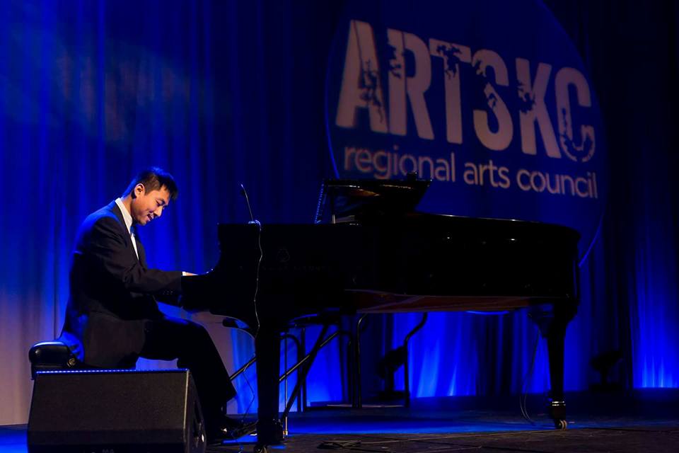 Person playing piano on stage with blue curtains and a banner reading 'ARTSC regional arts council'.